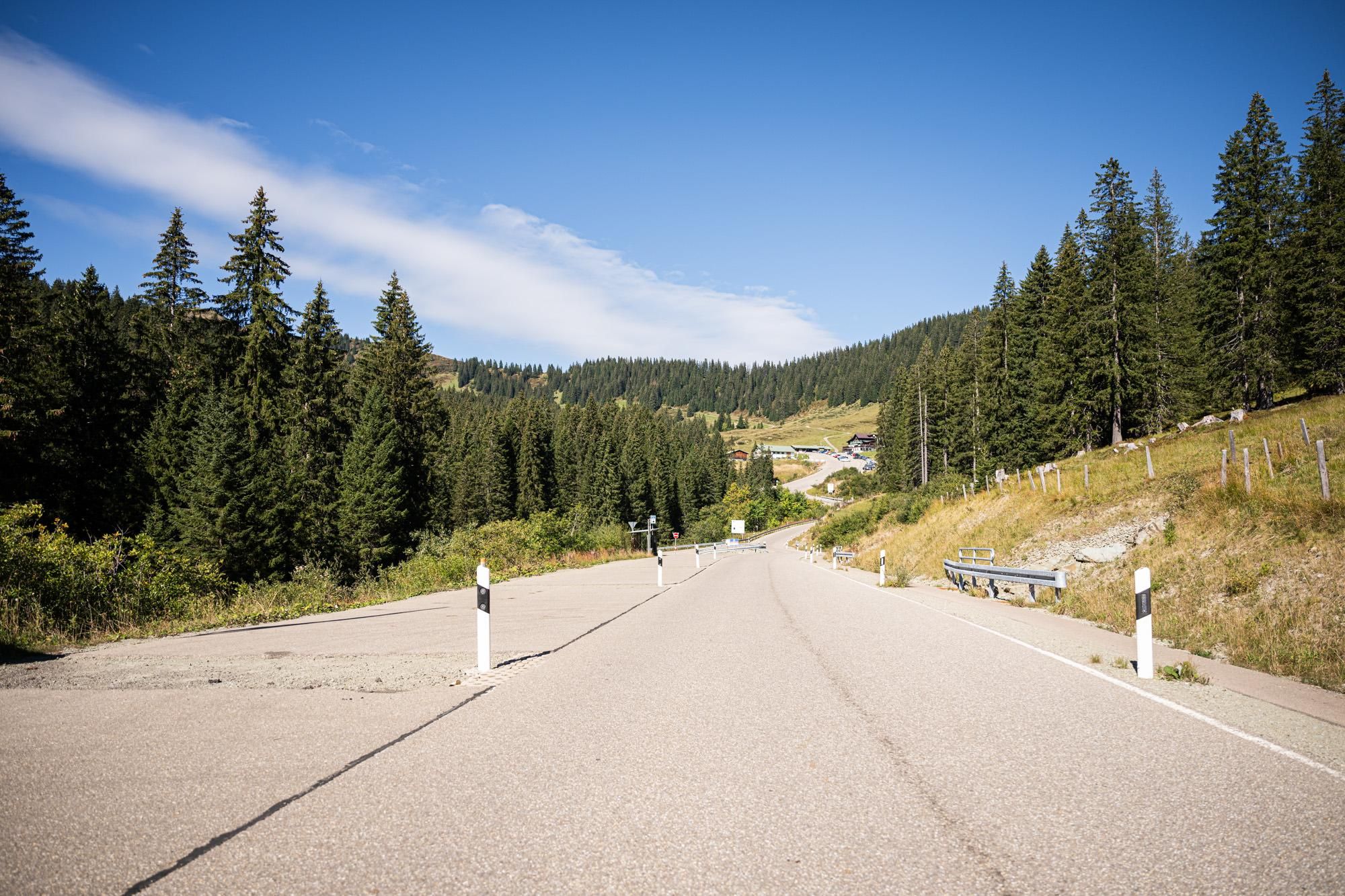 Asphaltierte Straße führt bergauf, umgeben von grünen Hängen mit Tannenbäumen, blauer Himmel mit Wolken.
