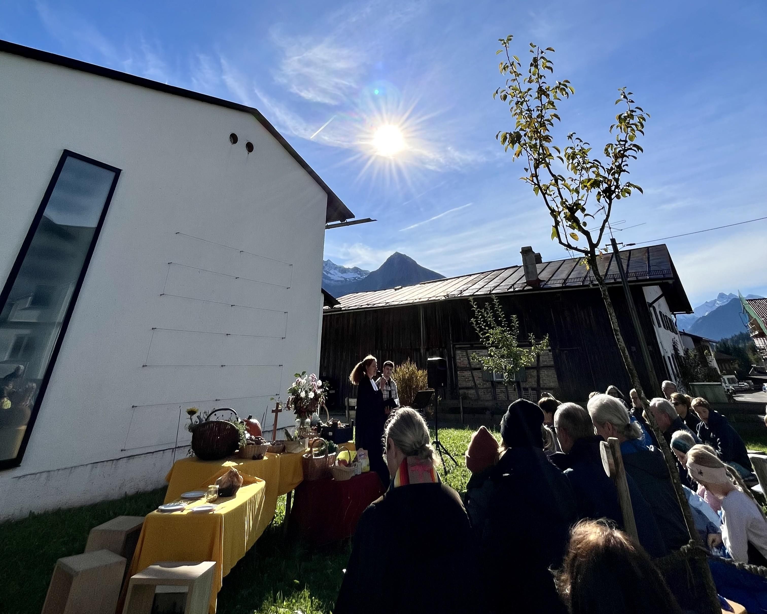 Die Gottesdienstbesucher sitzen hinter der Kirche auf einer Wiese. Der Himmel ist klar und die Sonne scheint hell.