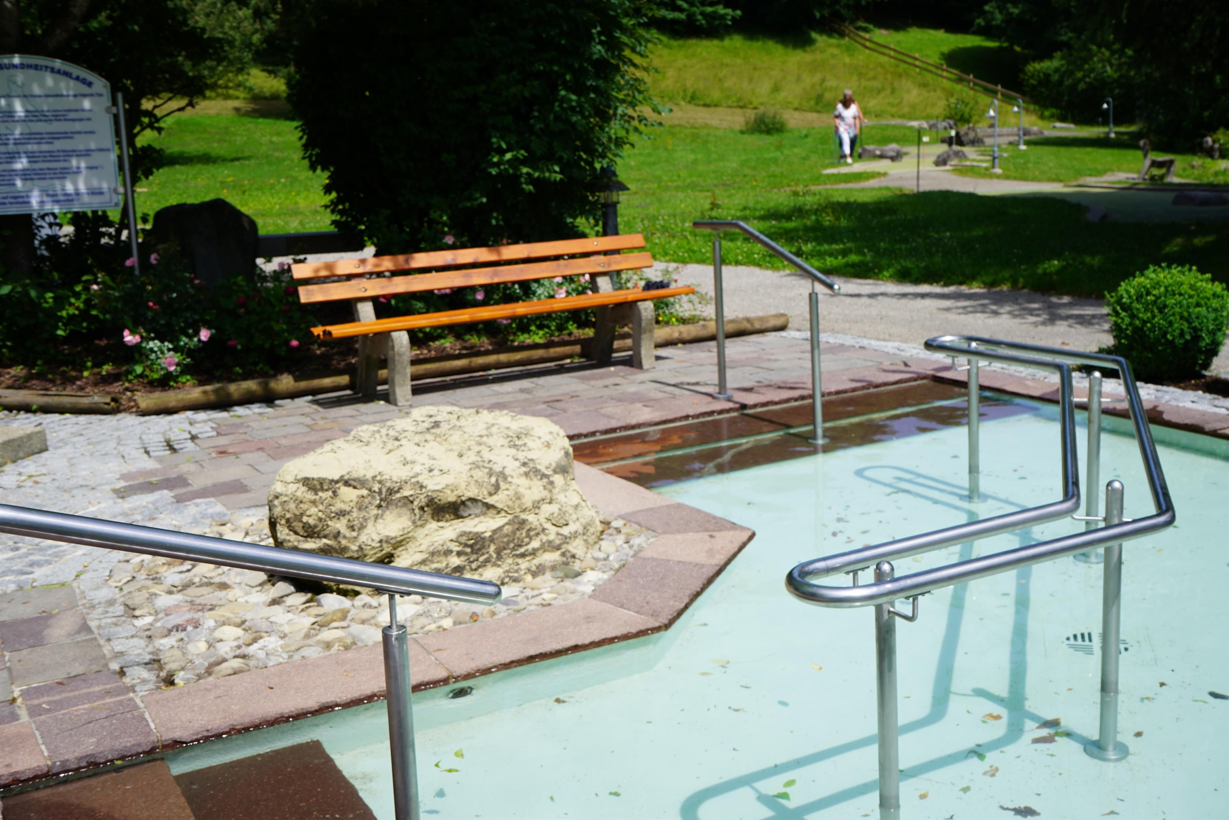 Kneippbecken mit hellem Wasser und silbernen Handläufen. Eine hölzerne Bank steht auf einem gepflasterten Bereich daneben. Grüne Wiese und Bäume im Hintergrund.