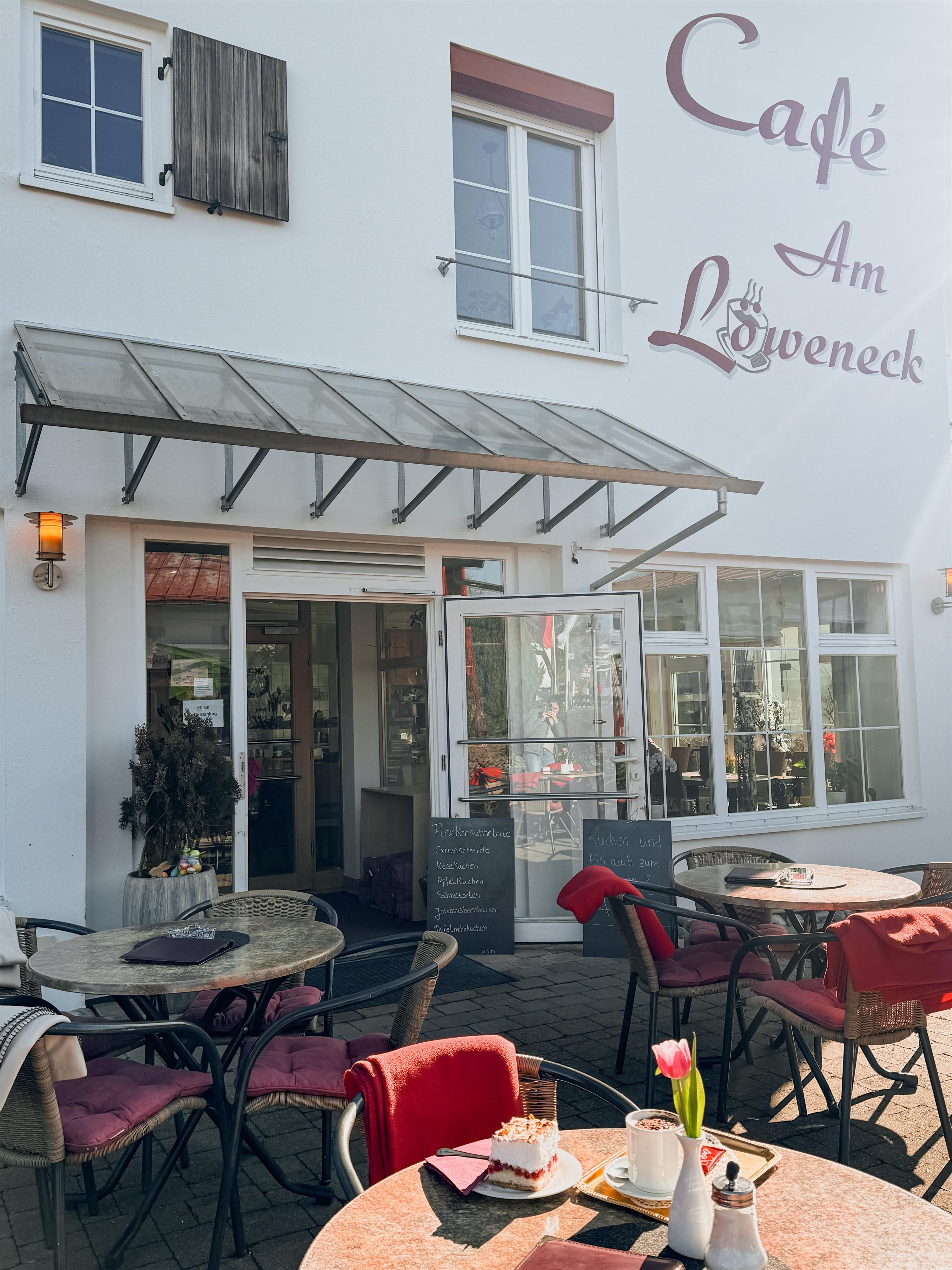 Außenansicht des Café Am Löweneck mit sonniger Terrasse, gedeckten Tischen und einladender Fassade.