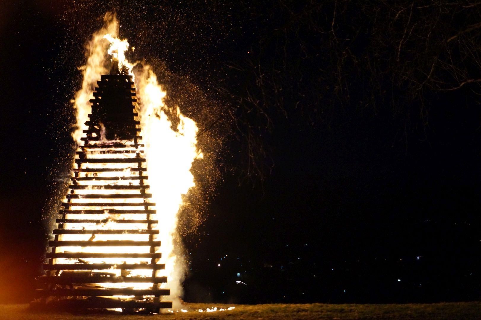 Gewaltiges Holzturm-Feuer in der Dunkelheit, Funken fliegen, Baumsilhouette rechts.