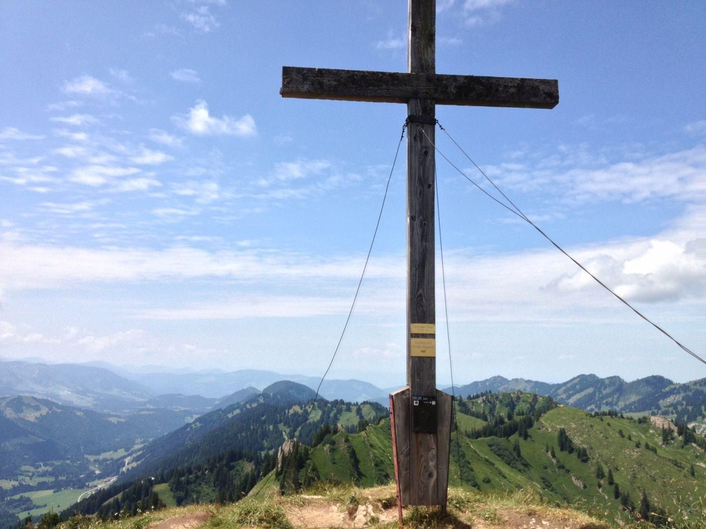 Hölzernes Gipfelkreuz auf Berg mit weitem Blick über grüne Täler und Berge.