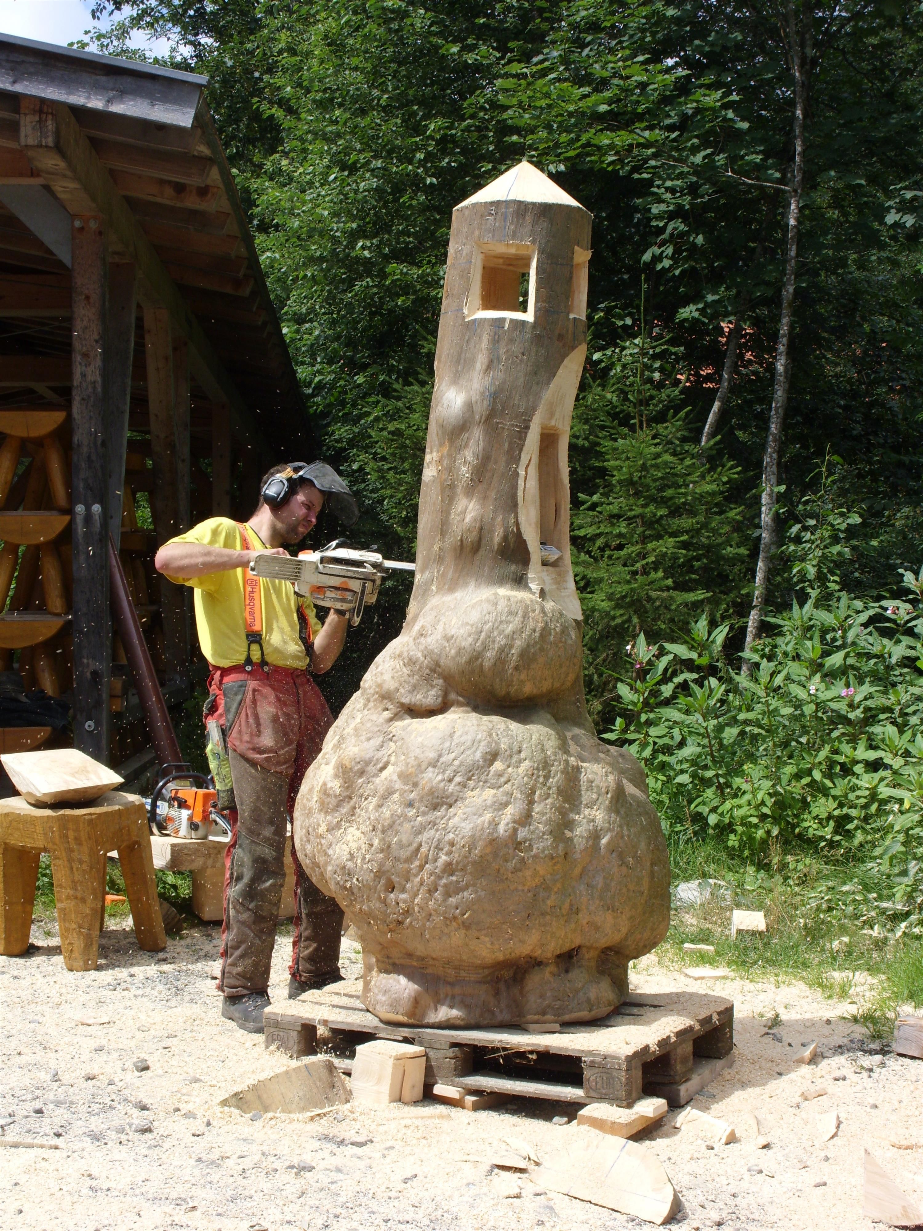 Ein Mann mit Helm und Schutzkleidung schnitzt mit einer Motorsäge eine Skulptur aus Holz im Freien.