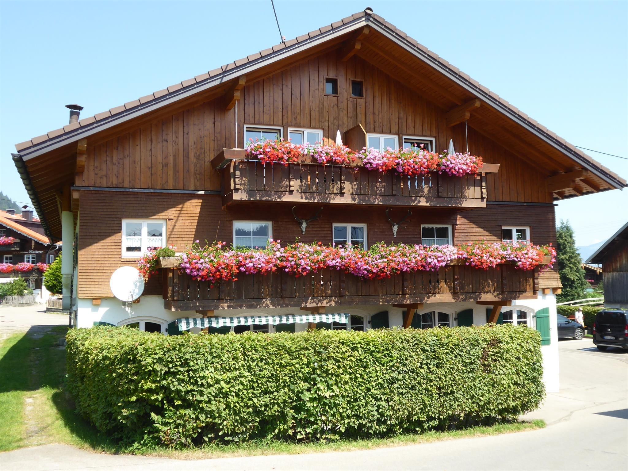Gasthof Riedbergerhorn in Grasgehren. Braunes Holzhaus mit zwei blumengeschmückten Balkonen. Grüne Hecke im Vordergrund.