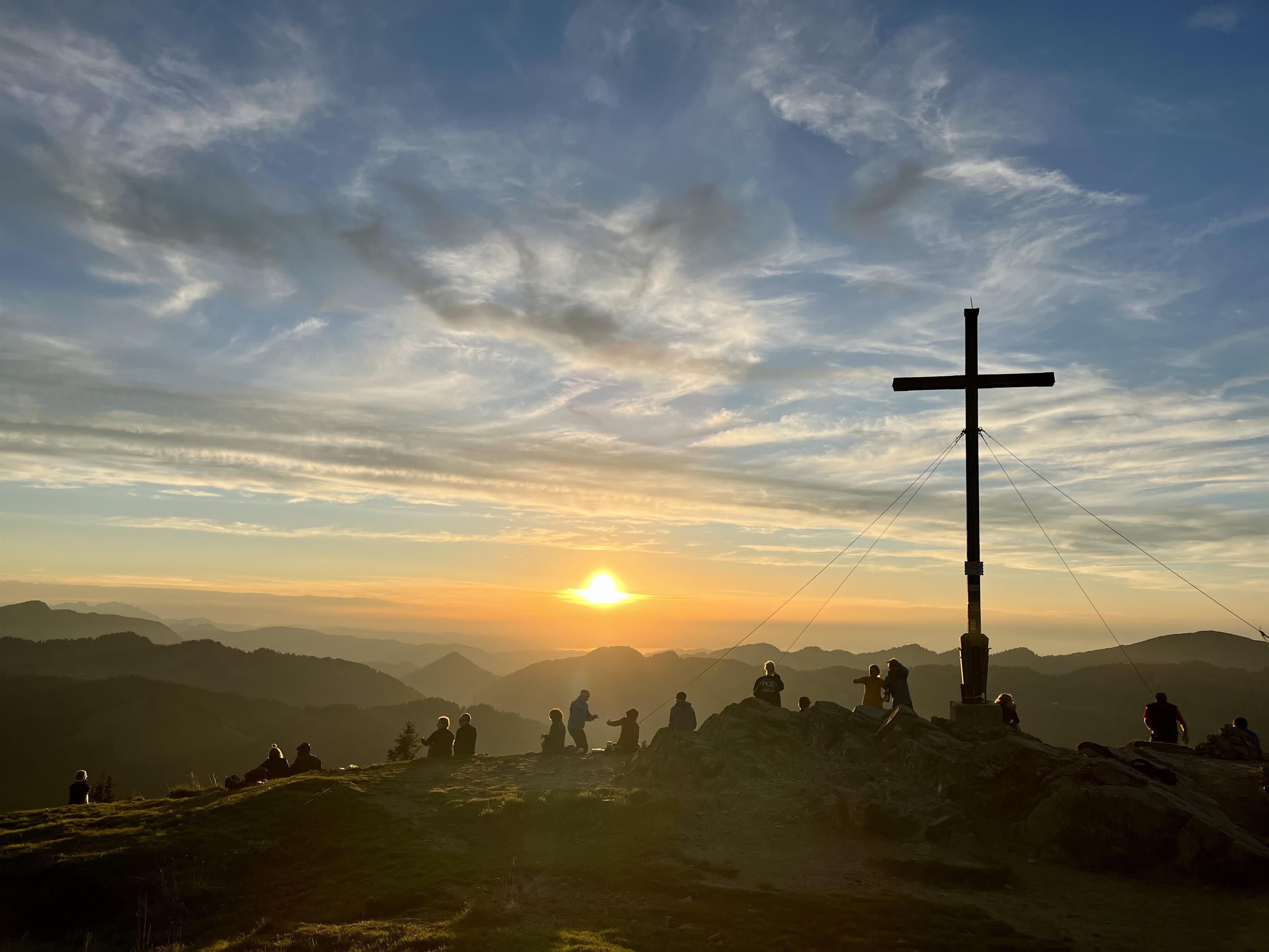Eine Gruppe von Menschen befindet sich rund um das Gipfelkreuz am Reidberger Horn, die Sonne geht bald unter.
