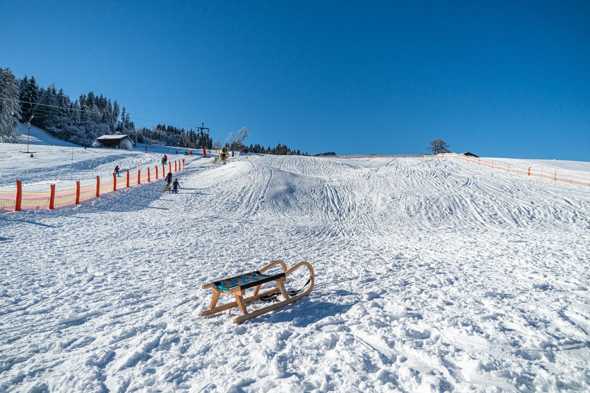 Ein Wintertag mit blauem Himmel, der Rodelhügel rechts neben dem Stinesser-Lift ist verschneit, vorne ein Schlitten.