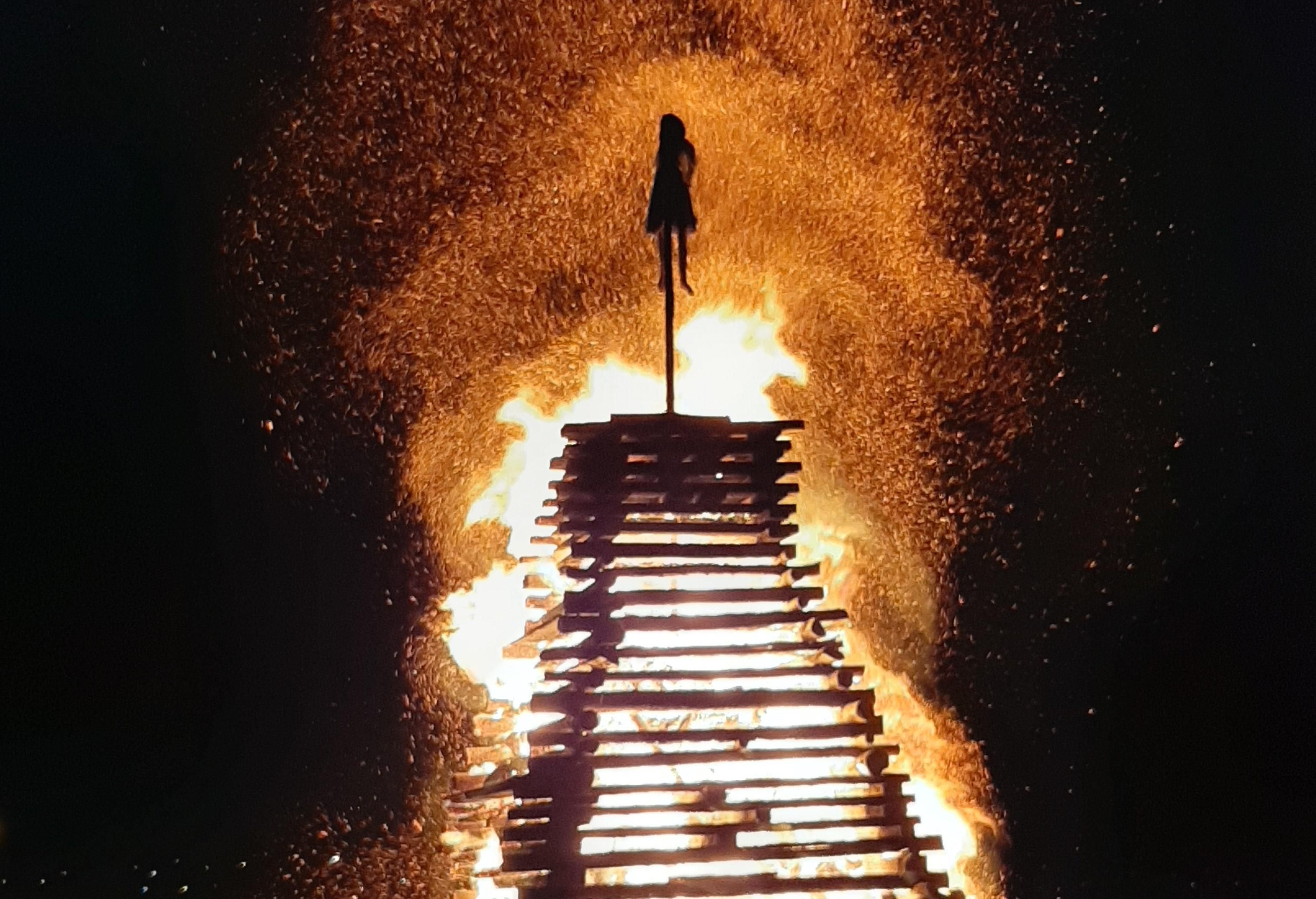 Holzscheit-Stapel brennt lichterloh, Funken sprühen, eine Figur auf einem Stock oben.