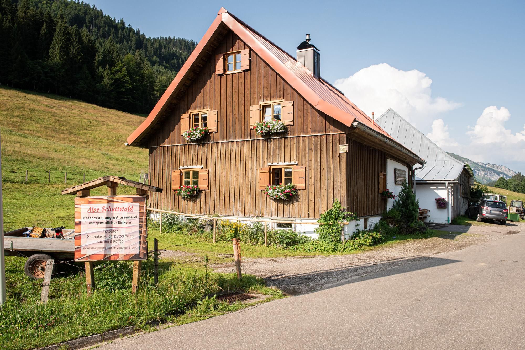 Eine traditionelle Holzalpe mit einem roten Satteldach und Blumenkästen an den Fenstern. Im Hintergrund sind grüne Wiesen und Berge zu sehen.
