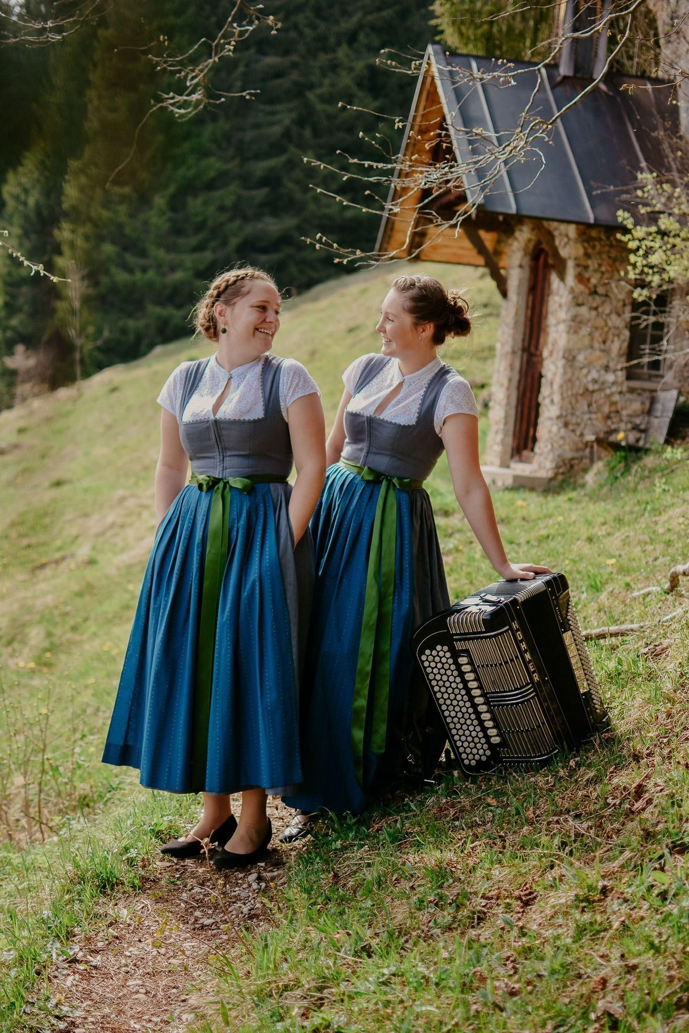 Zwei junge Frauen in Dirndl stehen lächelnd auf einem grasbewachsenen Hang. Eine hat ein Akkordeon bei sich. Im Hintergrund ein kleines steinernes Häuschen und Bäume.