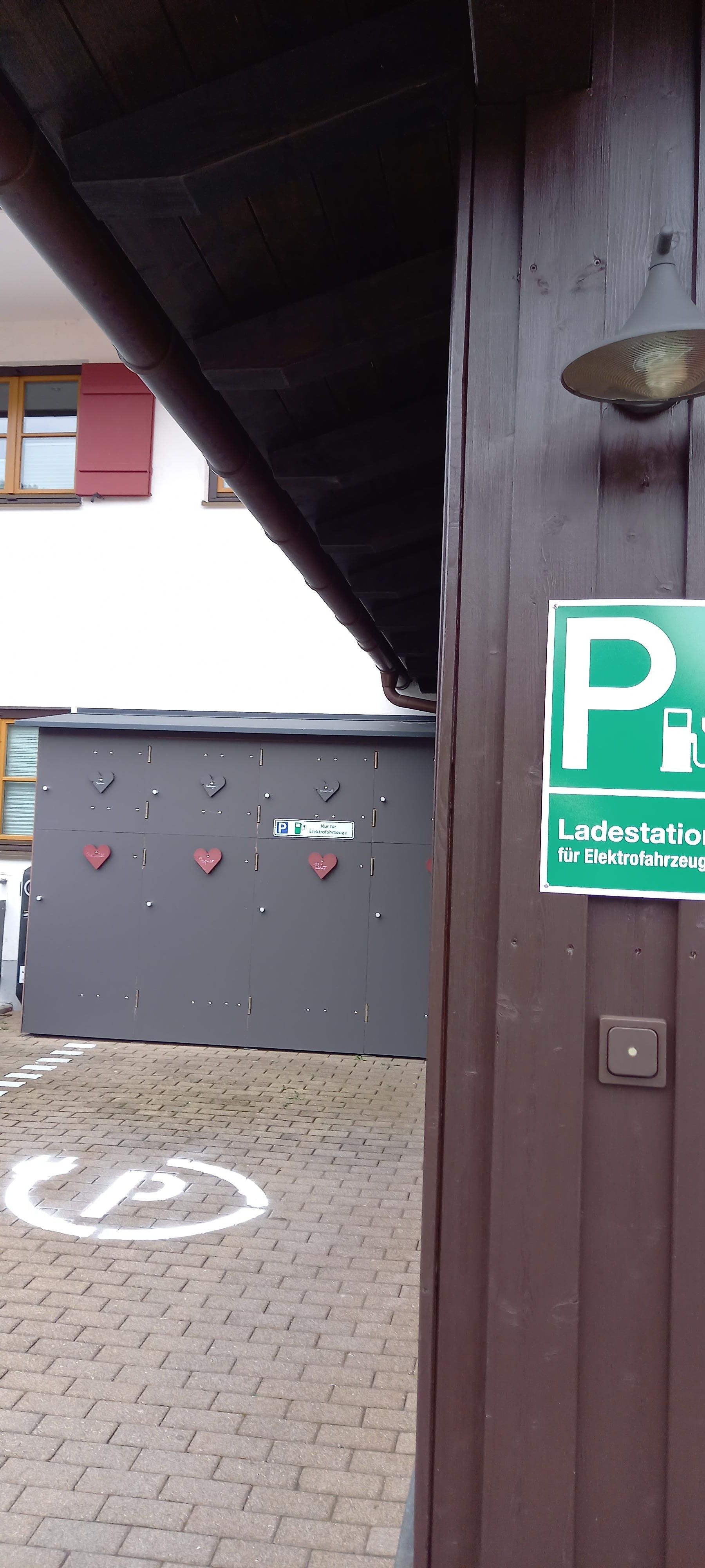 Ein Parkplatzschild mit grünem "P" und E-Ladesymbol an einer braunen Holzwand. Im Hintergrund ein grauer Kasten mit roten Herzsymbolen und ein gepflasterter Boden.