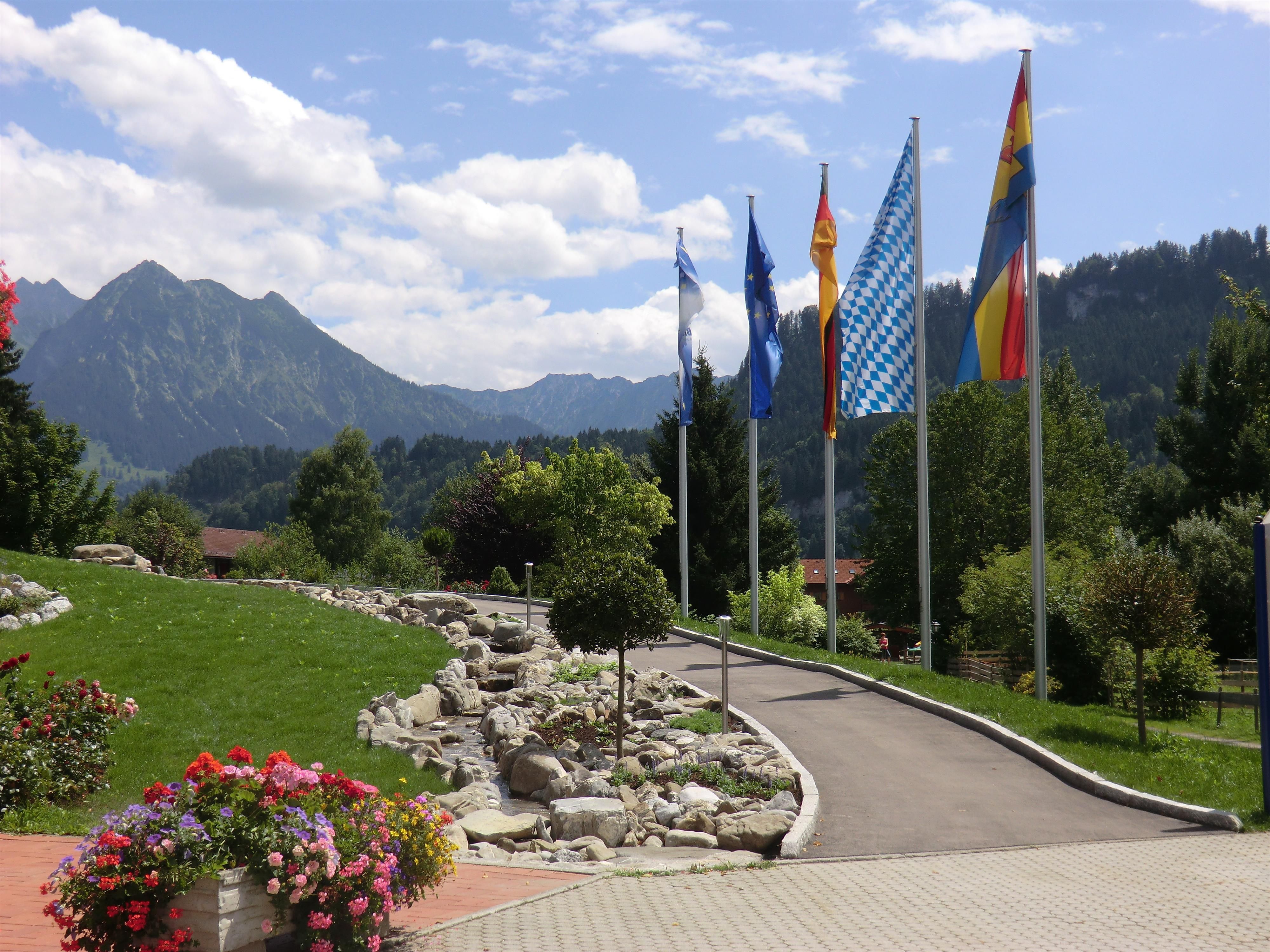 Gepflegter Außenbereich des Haus des Gastes Obermaiselstein. Ein gepflasterter Weg führt entlang von Fahnenmasten mit verschiedenen Flaggen.
