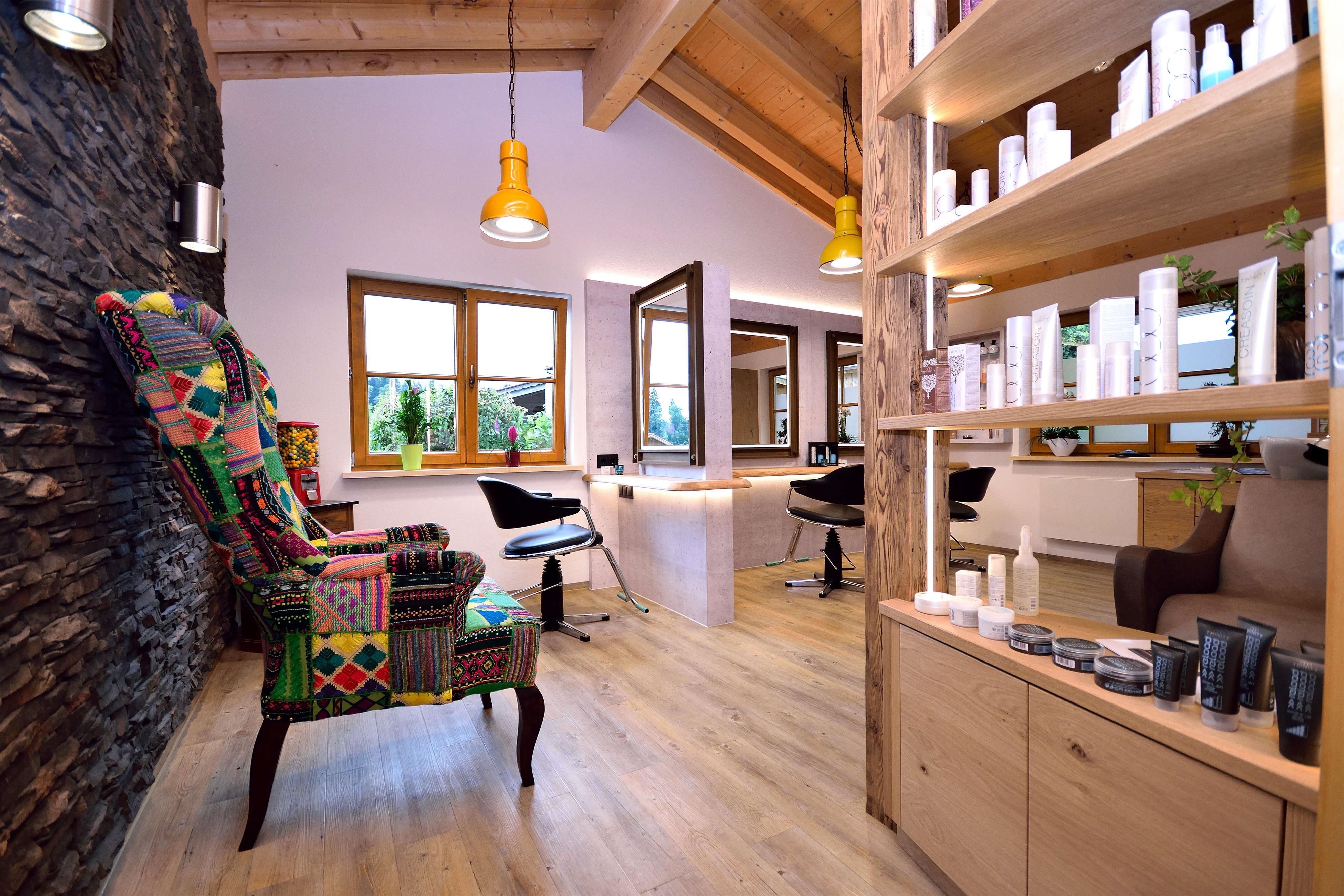 Friseursalon "Schneidig" mit Steinwand, Patchwork-Sessel, Frisierplätzen mit Spiegeln, Holzregalen voller Pflegeprodukte und Holzbalkendecke.