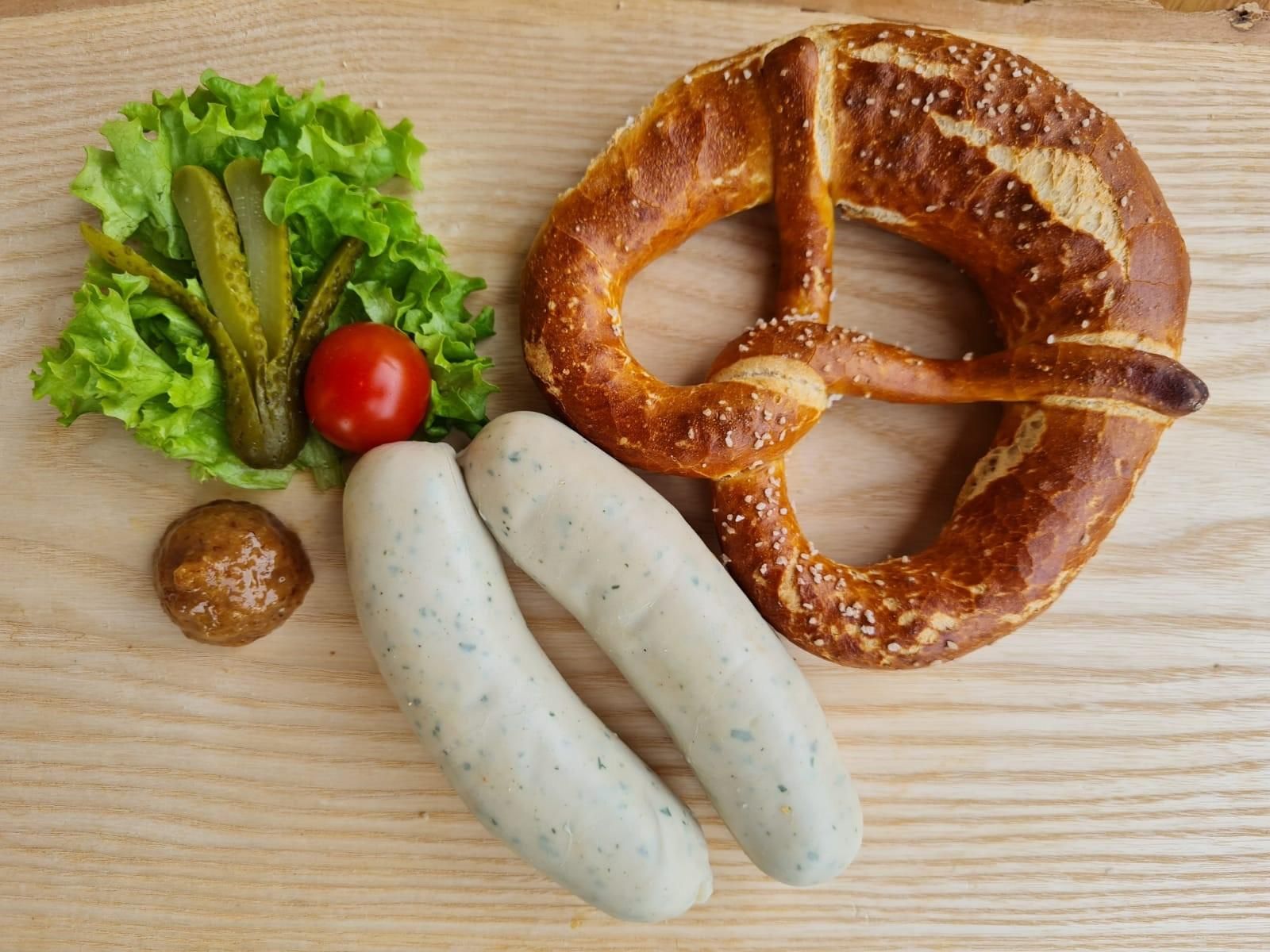 Zwei helle Weißwürste liegen diagonal auf einem Holzbrett. Daneben eine goldbraune Brezel mit grobem Salz.