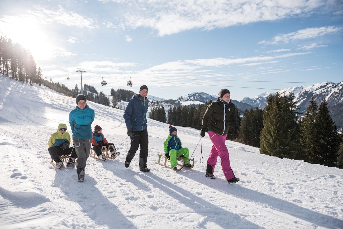 Eltern ziehen ihre Kidner auf Rodeln, im Hintergrund die Bergbahn Ofterschwang