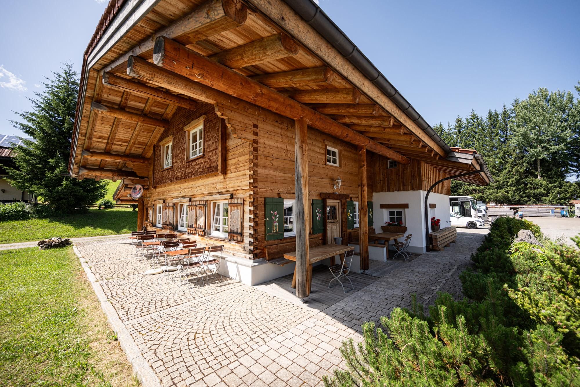 Gemütliches Holzgebäude des Wiesenstadls in Obermaiselstein. Außenbereich mit Tischen und Stühlen auf gepflastertem Untergrund.