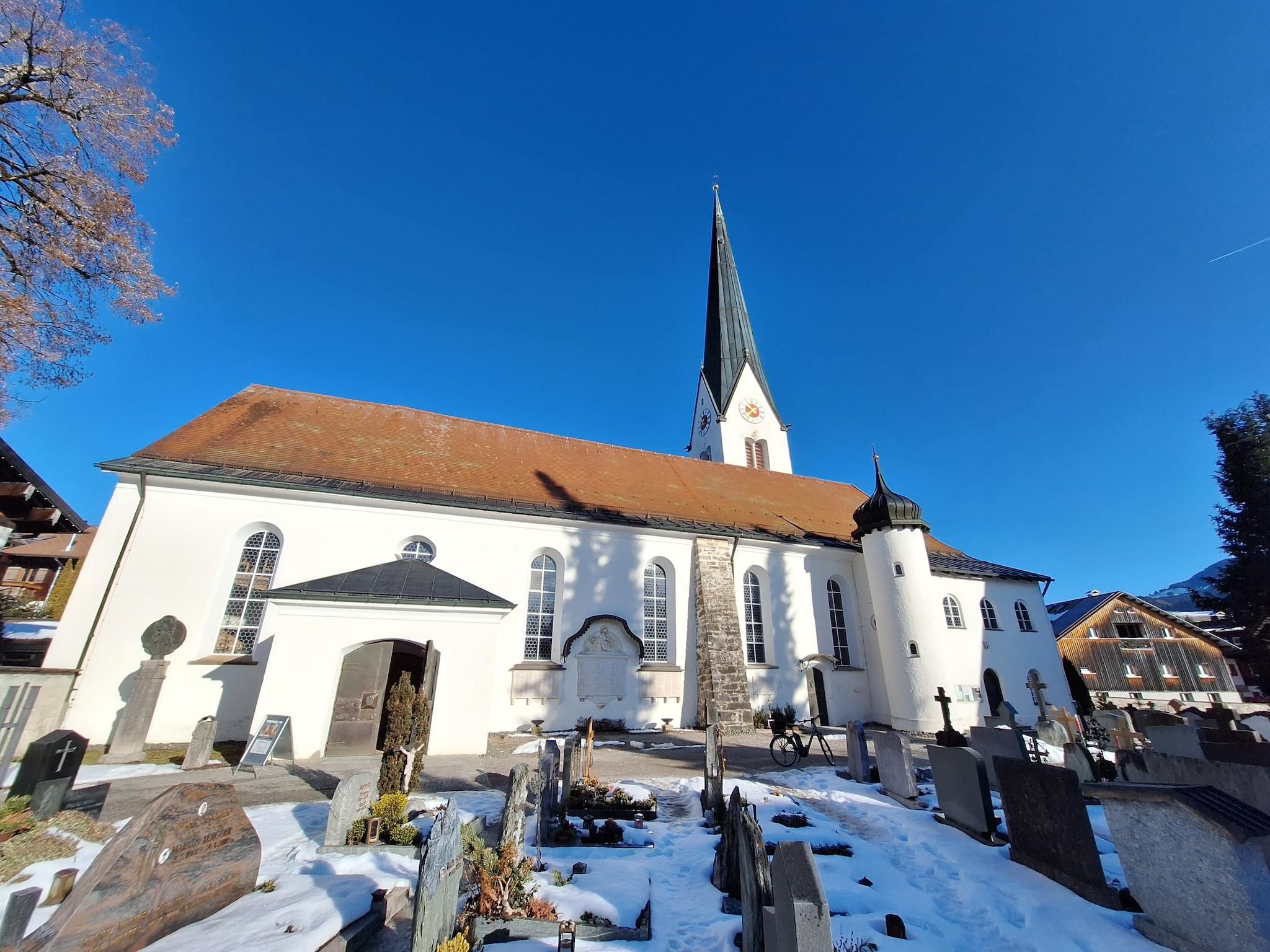 Eine Kirche mit einem hohen Turm und einem roten Ziegeldach. Im Vordergrund sind Gräber im Schnee zu sehen.