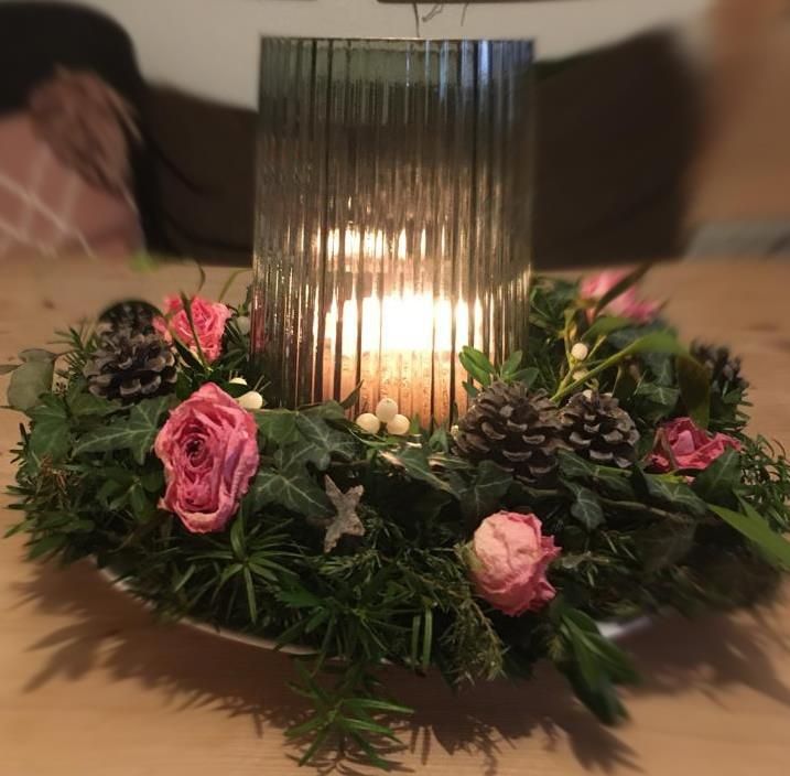 Ein Kranz aus Tannenzweigen mit kleinen Rosen in Pink und kleinen Zapfen, in der Mitte ein Glas mit Kerze.