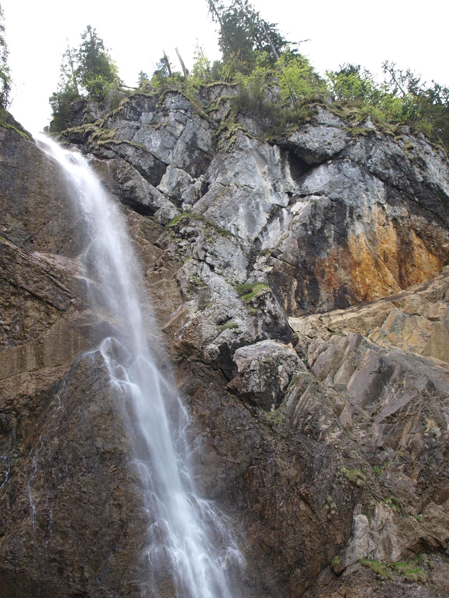 Ein hoher, schlanker Wasserfall stürzt eine steile, zerklüftete Felswand hinab. Die Felsen sind grau und braun gefärbt und weisen spärliche grüne Vegetation auf.