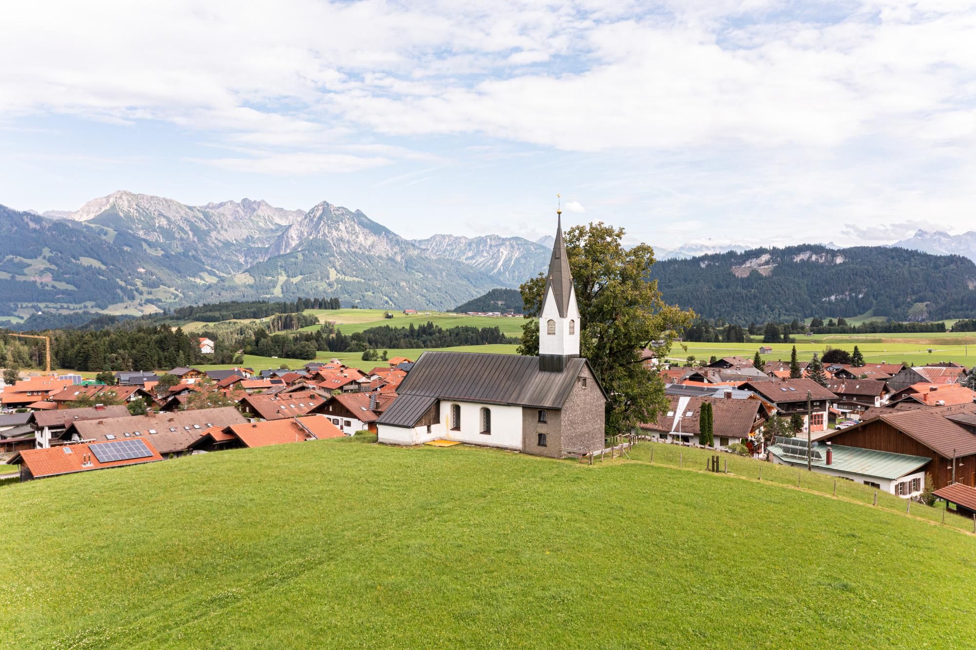 Kleine Kirche mit dunklem Dach und weißem Turm auf einem grünen Hügel. Umgeben von Häusern mit roten Dächern und Bergen im Hintergrund.