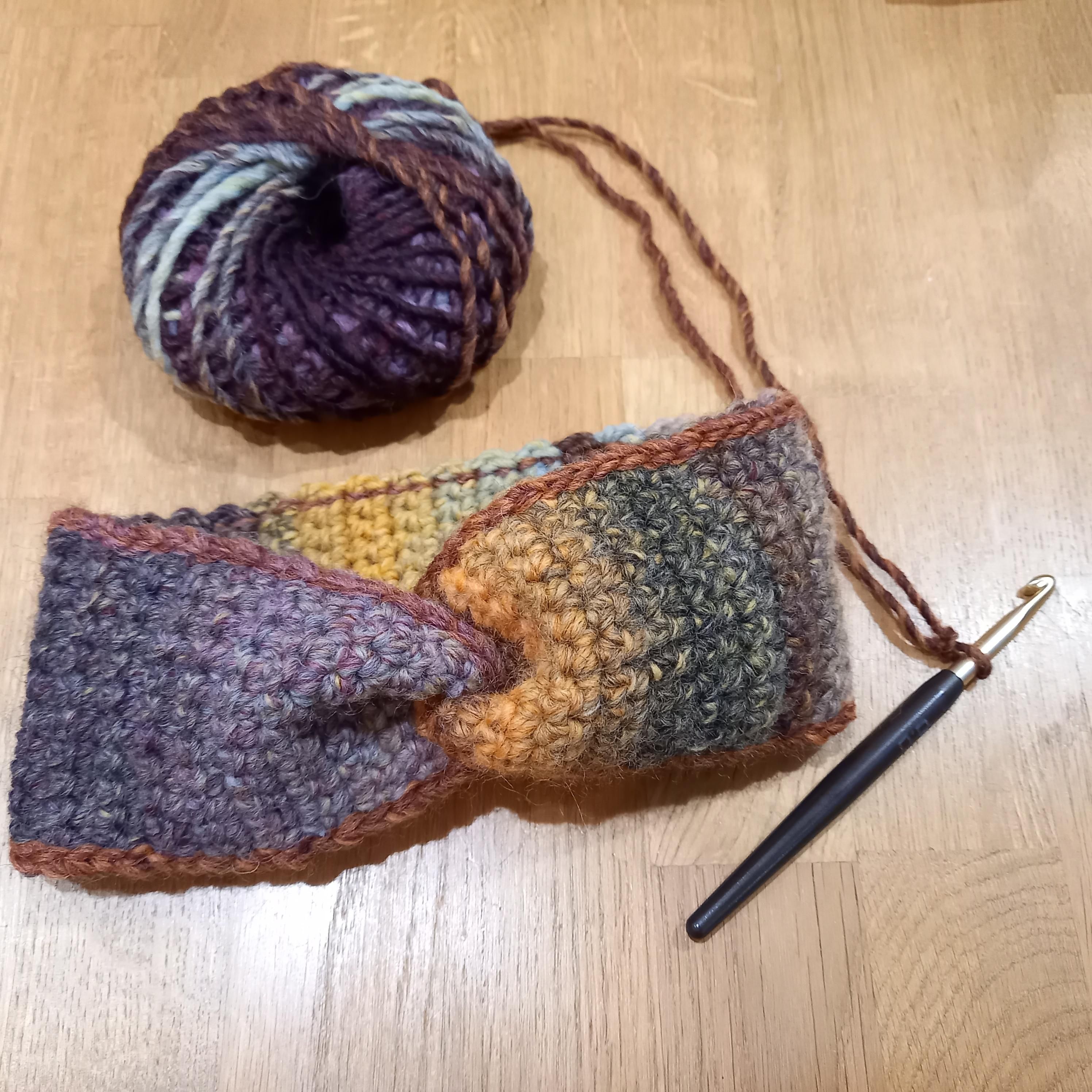 Ein Stirnband, die Wolle ist in verschiedenen Farben meliert, auf dem Foto liegt eine Häkelnadel neben dem Wollknäuel.