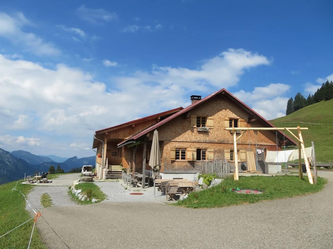 Blick auf die Piesenalpe. Vor der Holzhütte stehen Tische und Stühle sowie ein Sonnenschirm. Es gibt auch eine Schaukel.