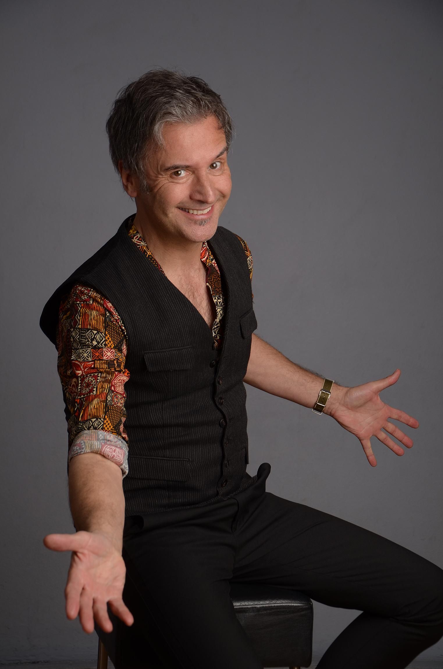 Chris Boettcher in Pose mit ausgebreiteten Armen bei seiner Kabarettshow gekleidet mit schwarzer Jeans, schwarzer Weste und rot gemustertem Hemd - halb sitzend, halb lehnend auf einem Barhocker.