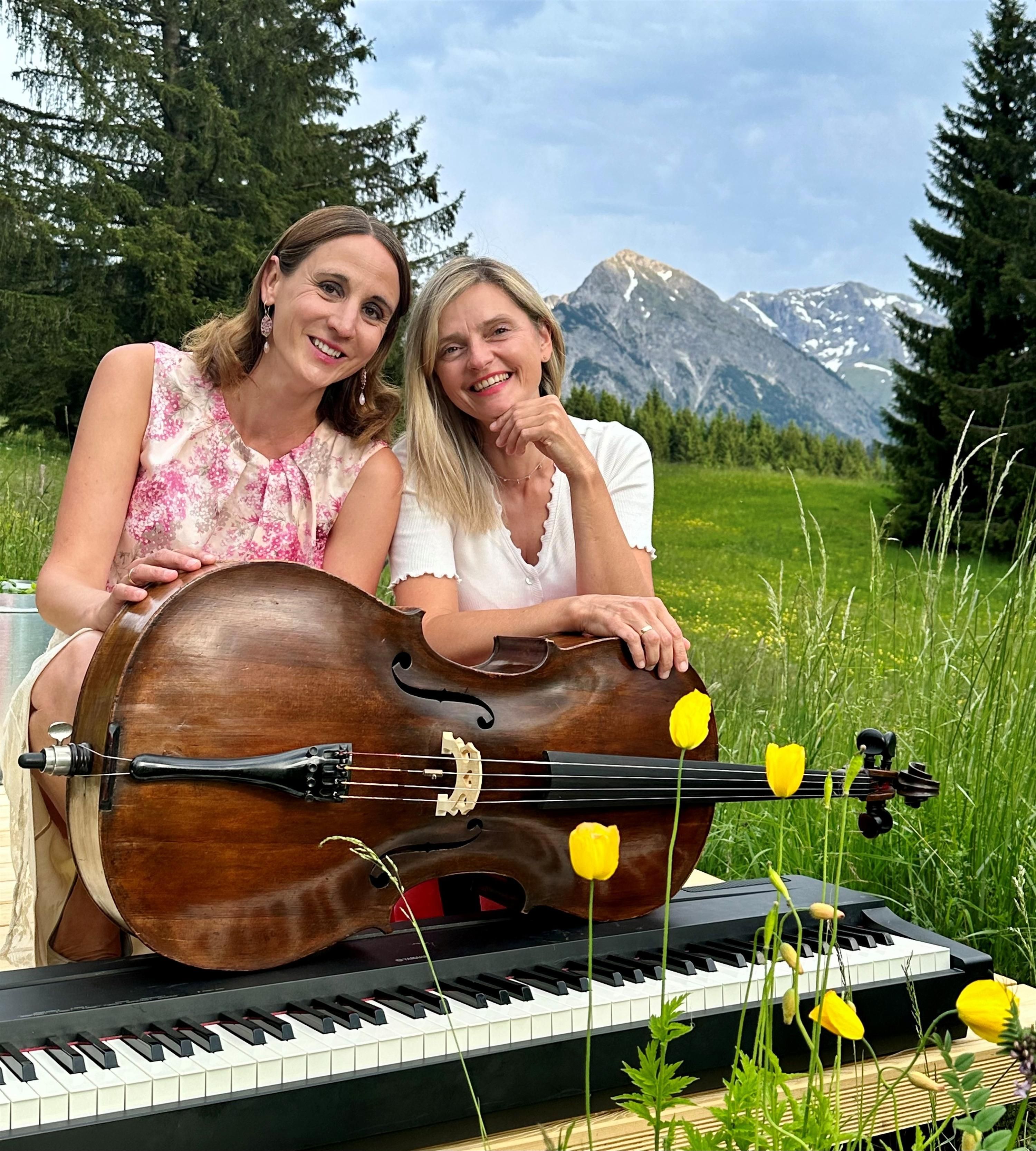 Zwei Frauen bilden das Duo Markato - hier abgebildet mit ihren Instrumenten Cello und Keyboard in einer saftig grünen Wiese vor Bergkulisse und Tannen.