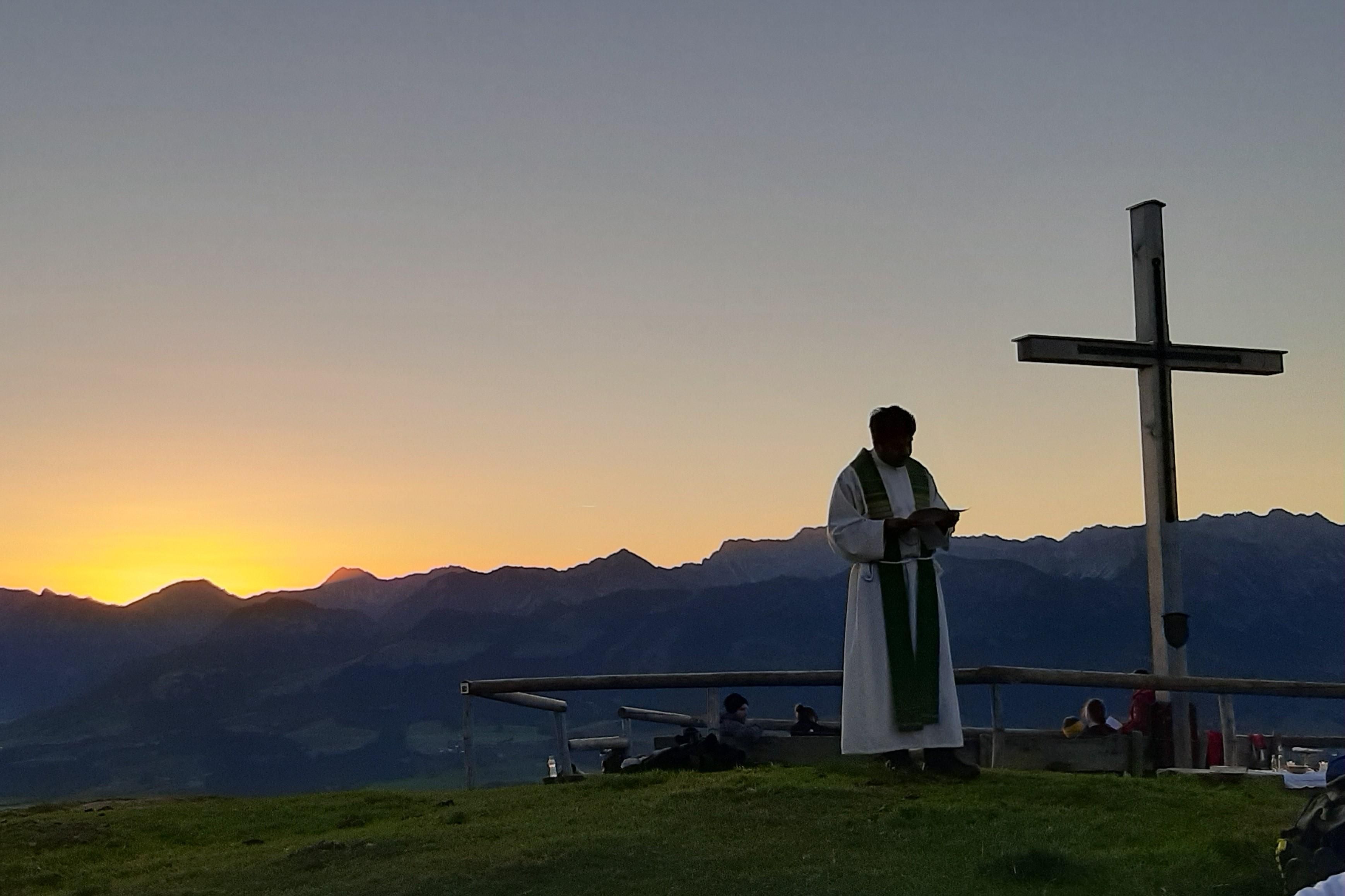 Auf dem Ofterschwanger Horn findet eine Bergmesse statt. Ein Priester steht vor einem Holzkreuz bei Sonnenuntergang.