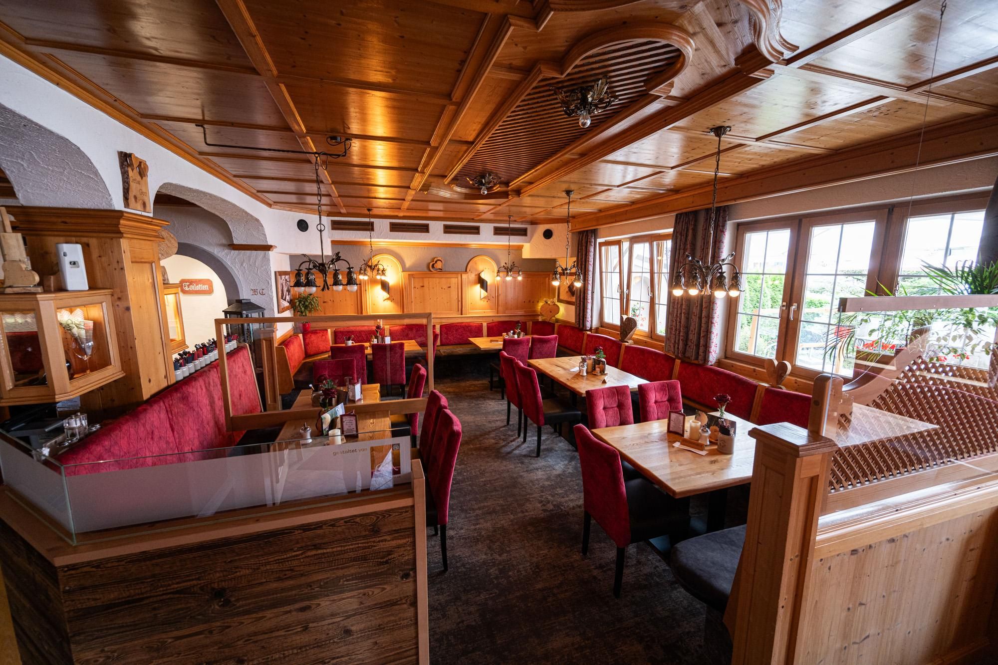Holzvertäfelte Decke und Wände. Rote Polsterbänke und Stühle an hellen Holztischen. Fenster mit gemusterten Vorhängen.