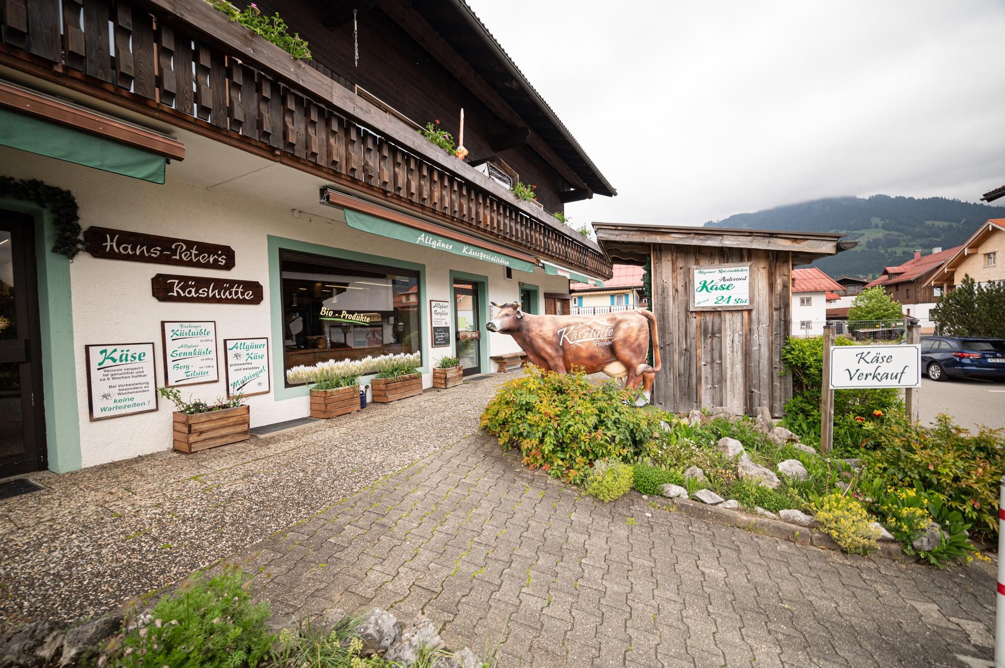 Käshütte in Fischen, links der Eingang zum Laden und ein großes Fenster, rechts eine Kuh vor dem Käseautomaten.