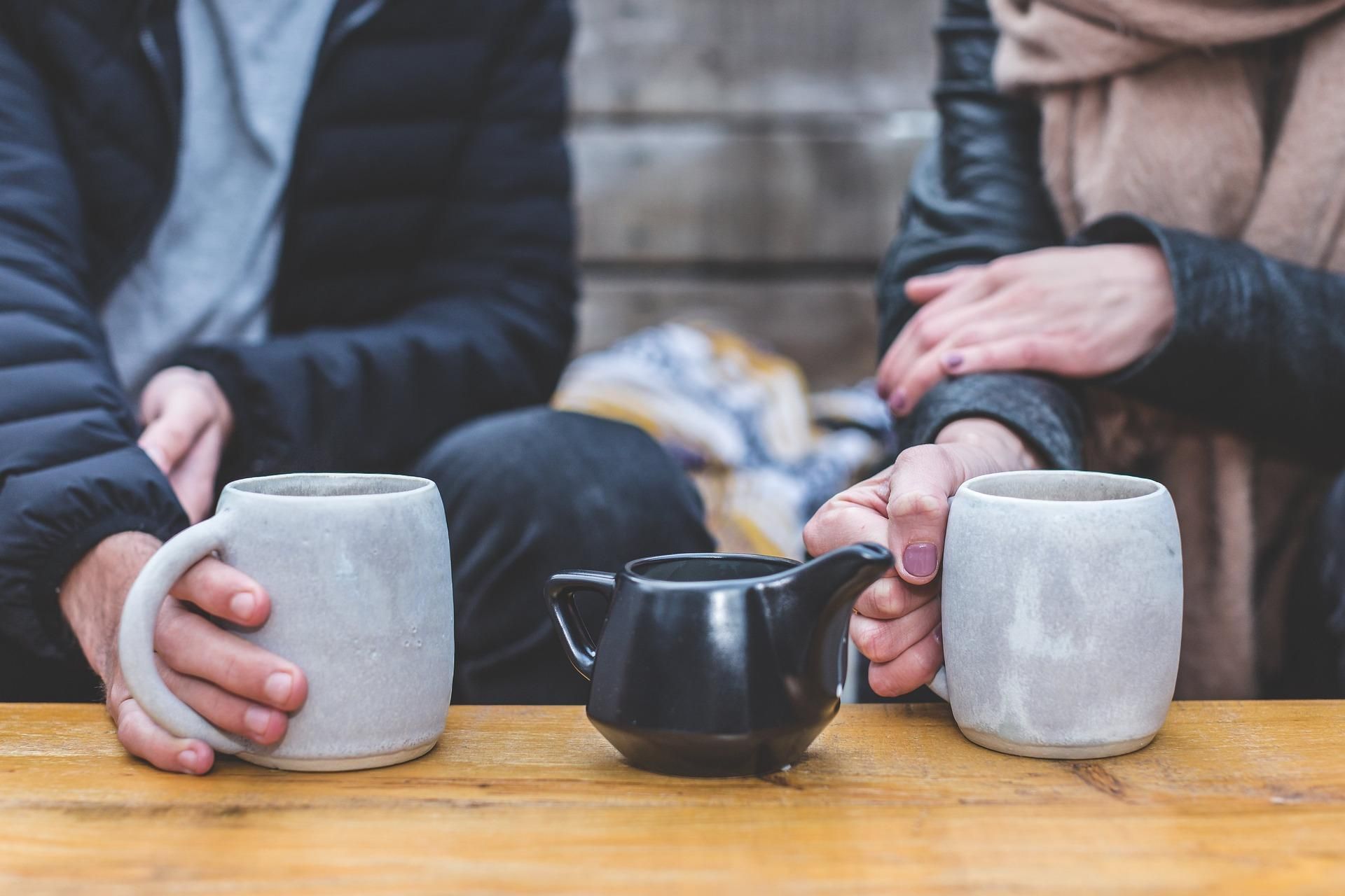 Zwei Personen sitzen an einem Couchtisch, zwei große, weiße Teebecher und eine schwarze Teekenne stehen auf dem Tisch.