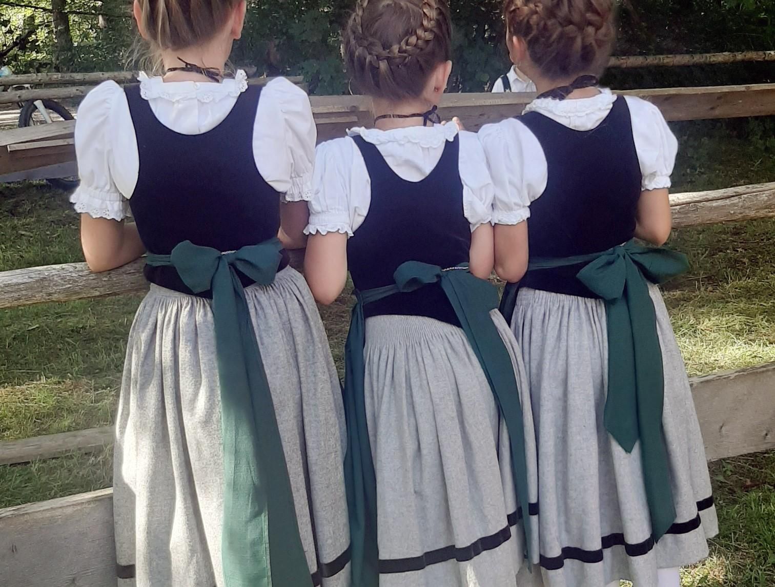 Drei Mädchen stehen an einem Zaun, ihre Dirndlkleider haben schwarze Oberteile und graue Röcke, die Schürzen sind grün.