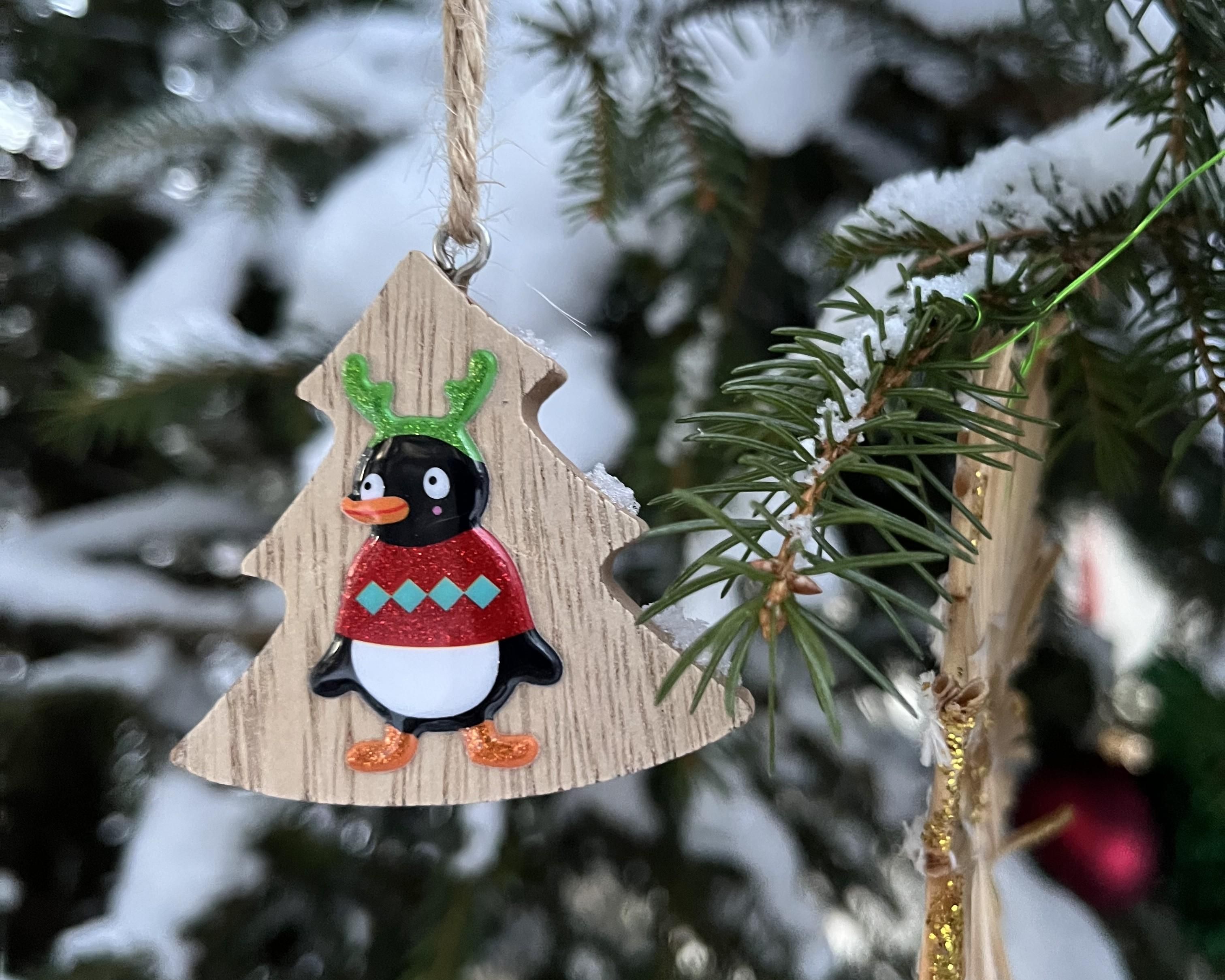 Ein Weihnachtsbaumanhänger in Form einer Tanne. Darauf abgebildet ist ein Pinguin, der einen bunten Pullover trägt. Der Anhänger hängt vor einem schneebedeckten Hintergrund.