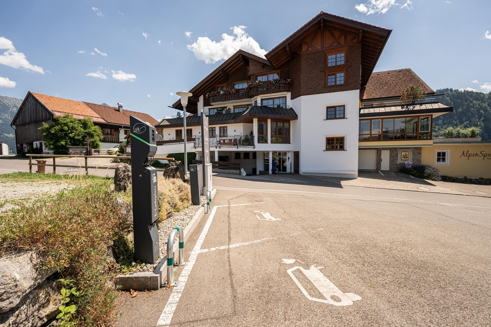 Eine E-Auto-Ladestation mit zwei Säulen vor einem hellen, mehrstöckigen Gebäude mit Holzelementen und Balkonen. Parkplätze mit E-Ladesymbolen sind markiert.