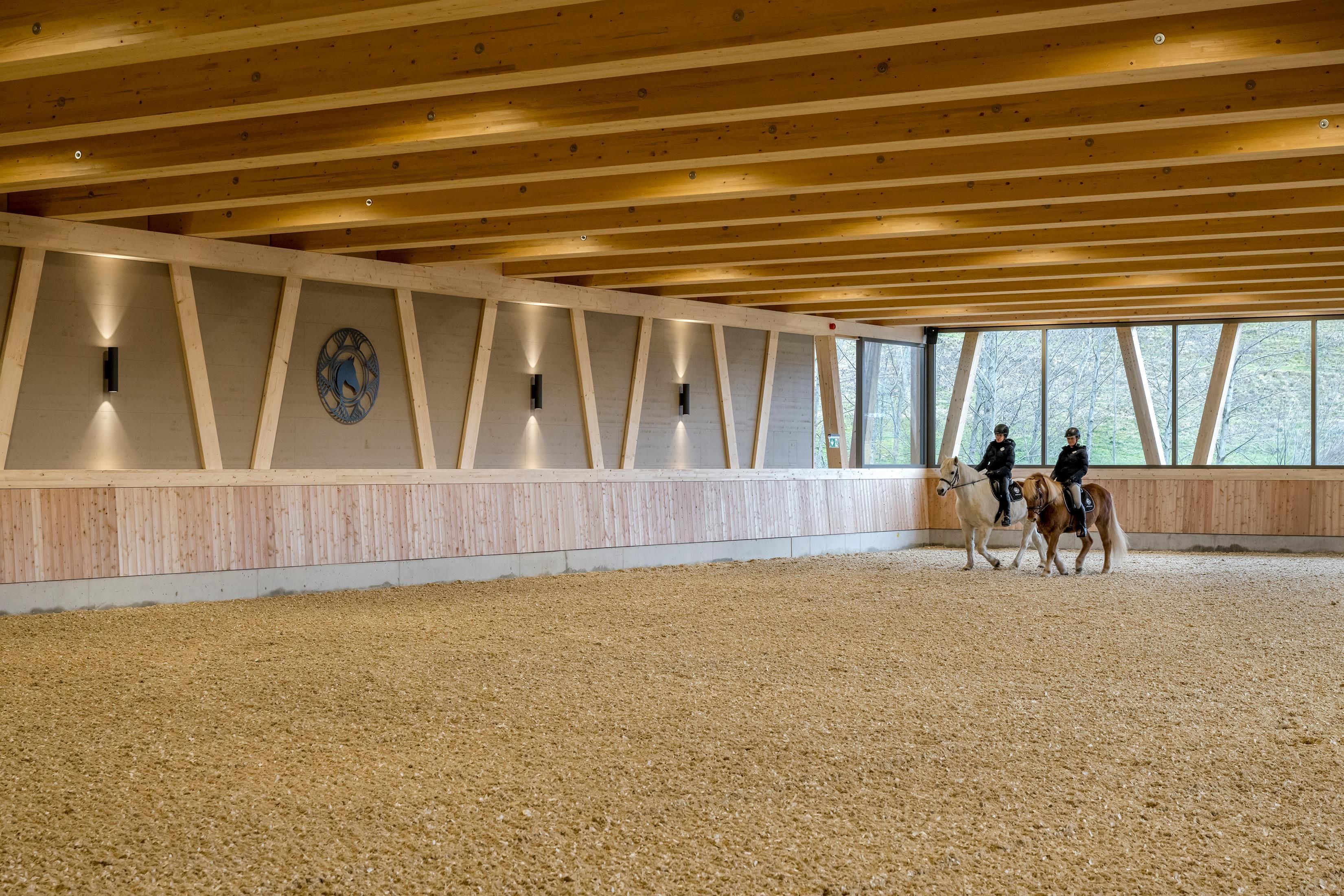 Eine Reithalle mit Holzdecken und großen Fenstern. Zwei Reiter auf Pferden üben in der Arena.