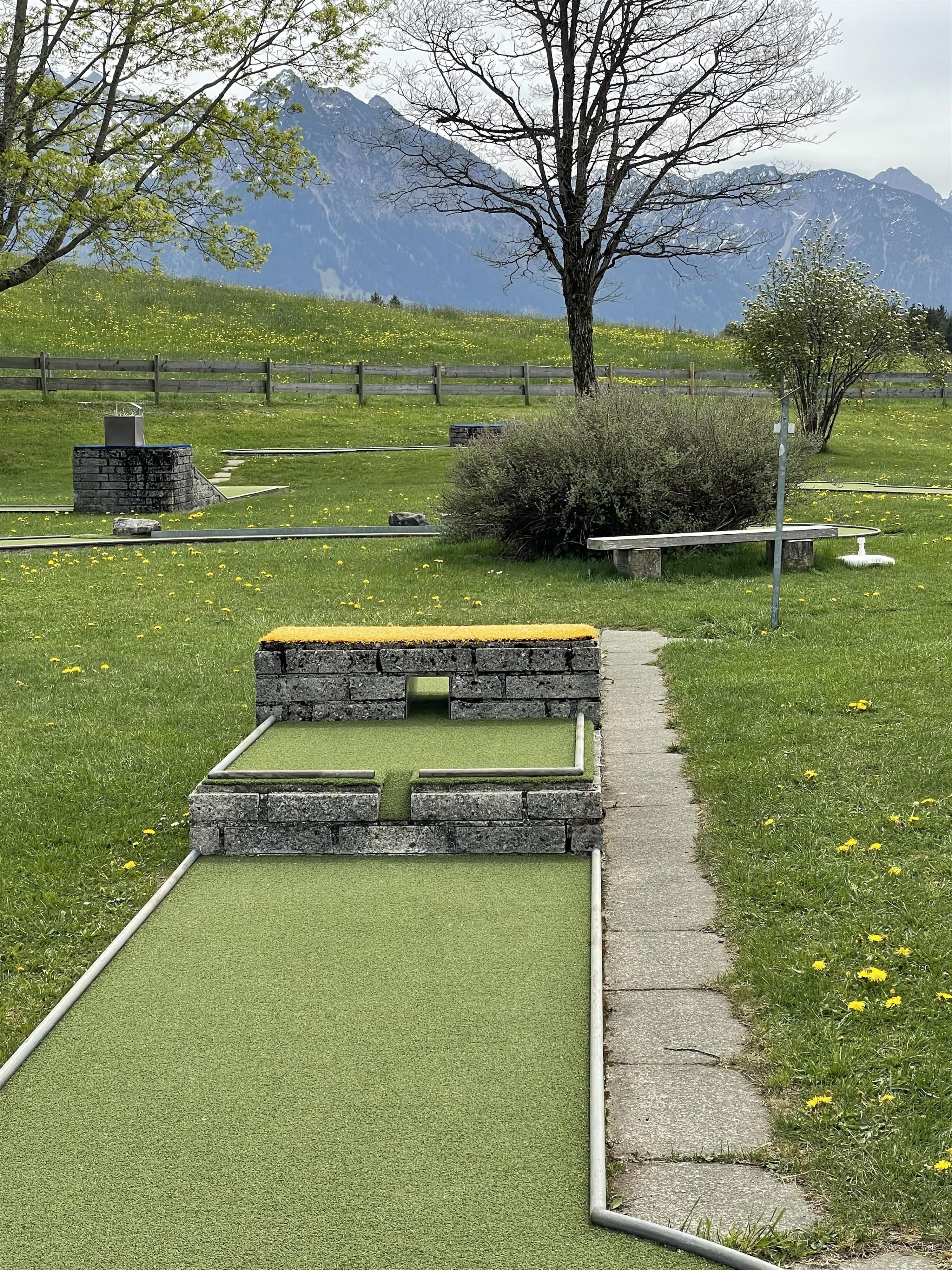 Ein Minigolfplatz mit einer künstlichen grünen Bahn und einer Mauer aus Steinen. Im Hintergrund sind Bäume und Berge zu sehen.