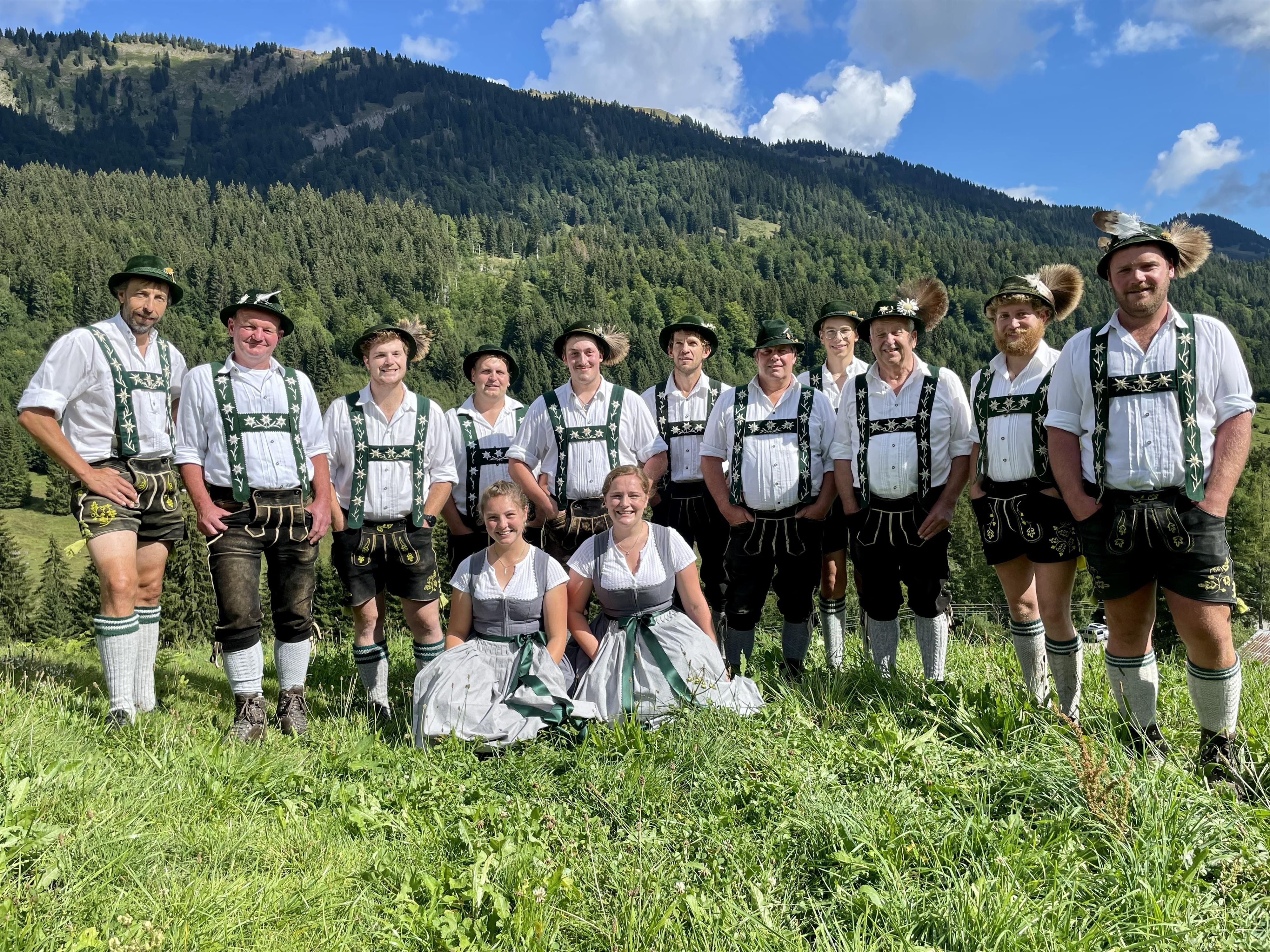 Eine traditionelle Jodlergruppe in bayerischer Tracht steht auf einer grünen Wiese vor einem bewaldeten Berghang.