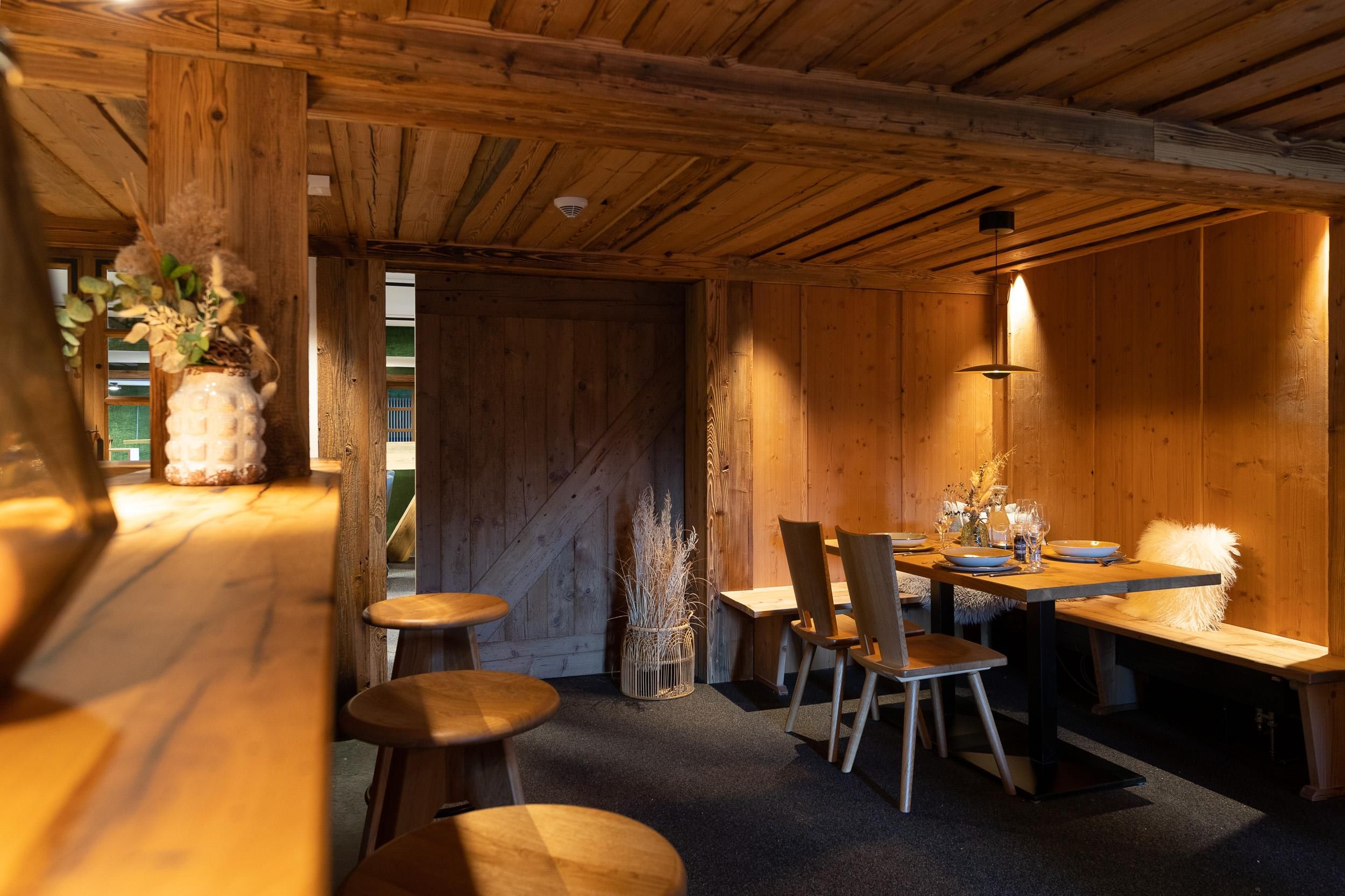 Rustikaler Innenraum der Köpfle Alpe mit Holzbalken und -wänden. Essbereich mit Holztisch, Stühlen und Bank. Barhocker im Vordergrund.