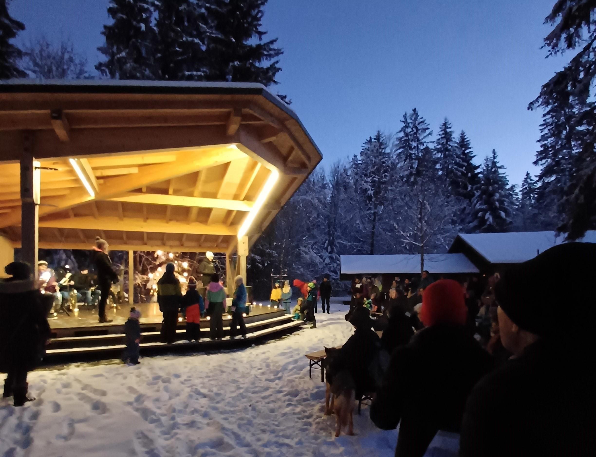 Ein festlicher Gottesdienst im Winter mit Menschen, die vor einer Holzbühne stehen. Schnee bedeckt den Boden und es gibt Bäume im Hintergrund.