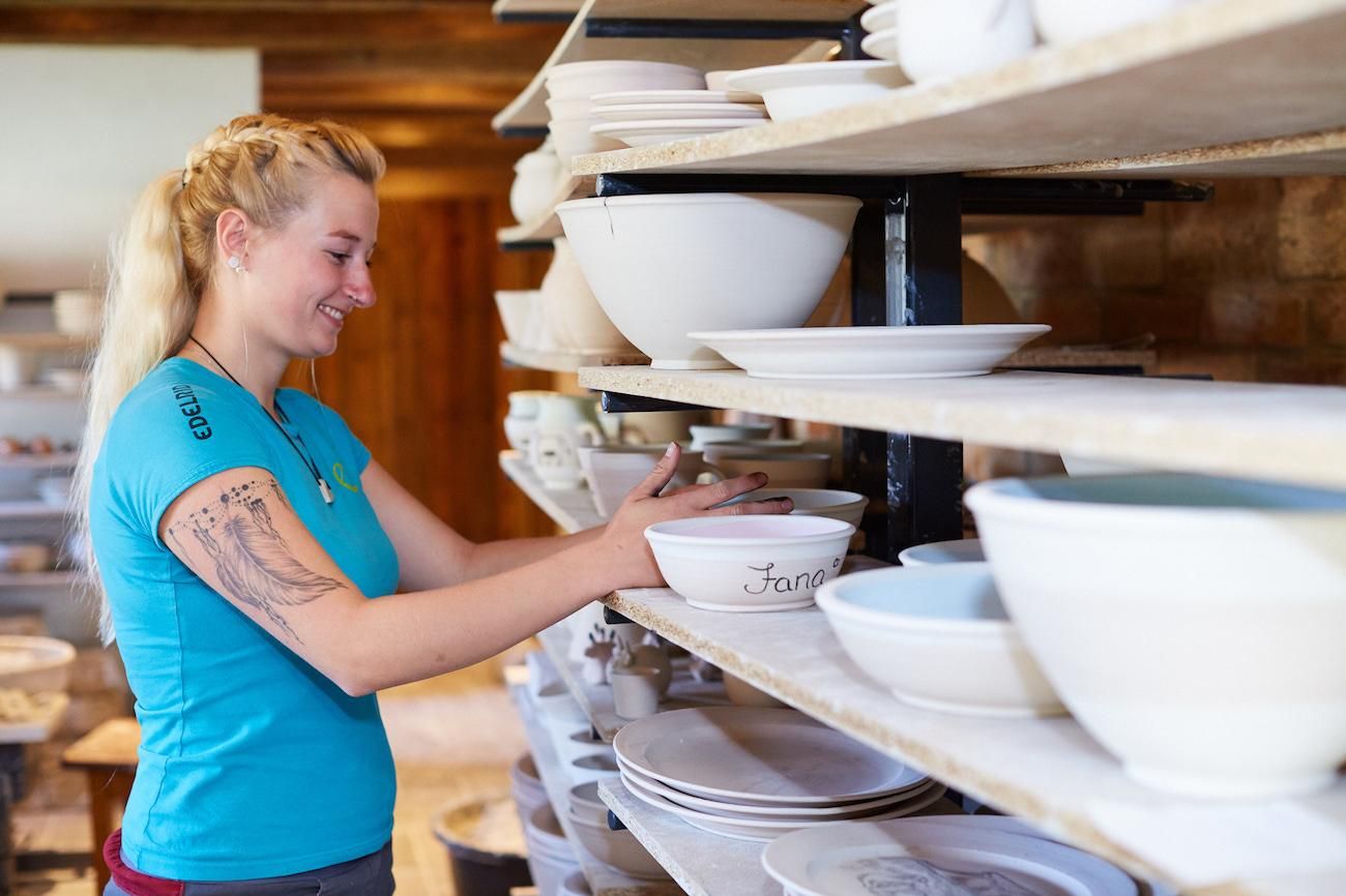 Frau mit blondem Zopf in einer Töpferei. Sie räumt weiße Keramikschalen in ein Regal.