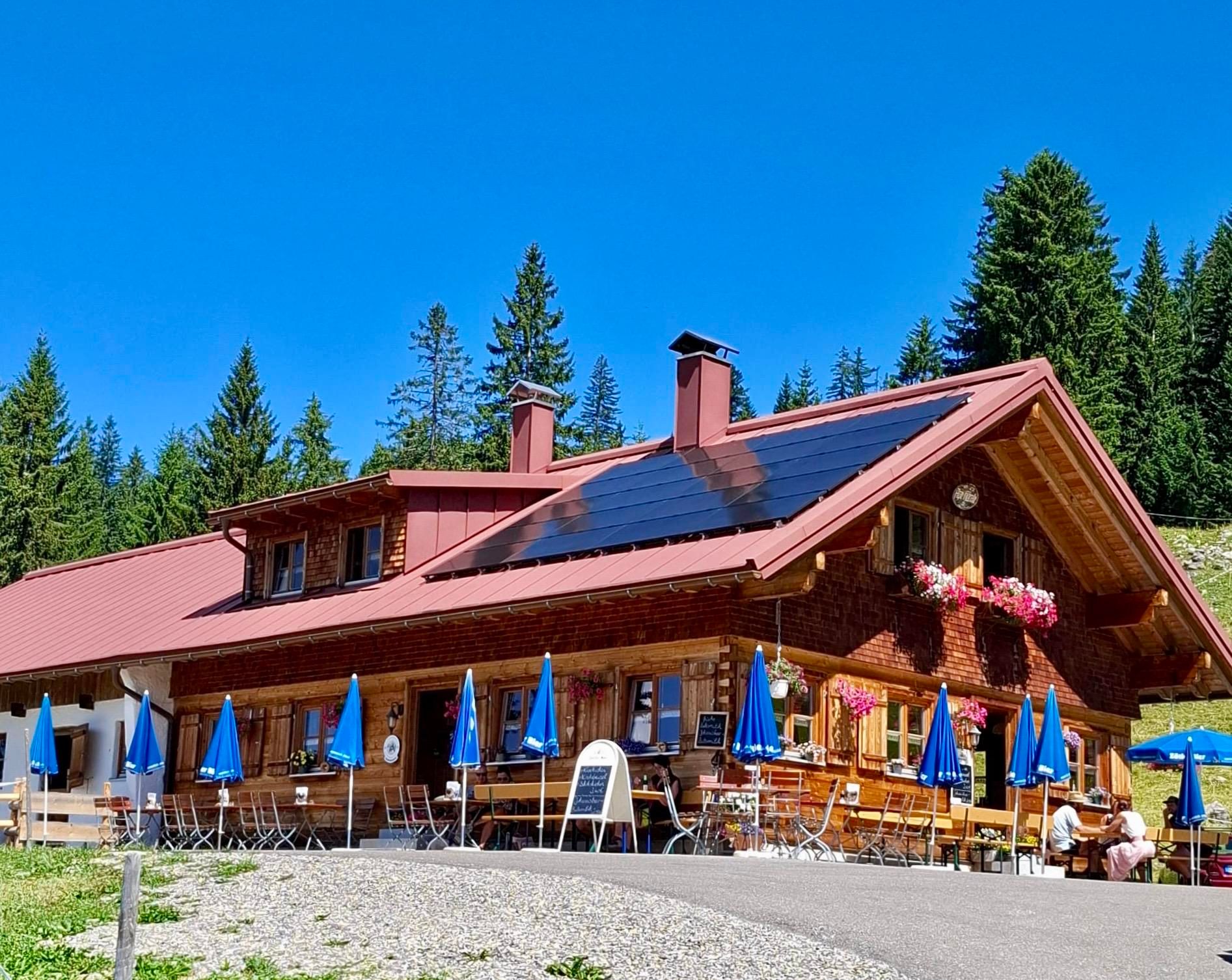 Eine gemütliche Alpe umgeben von Bäumen. Vor dem Haus stehen Tische mit blauen Sonnenschirmen.