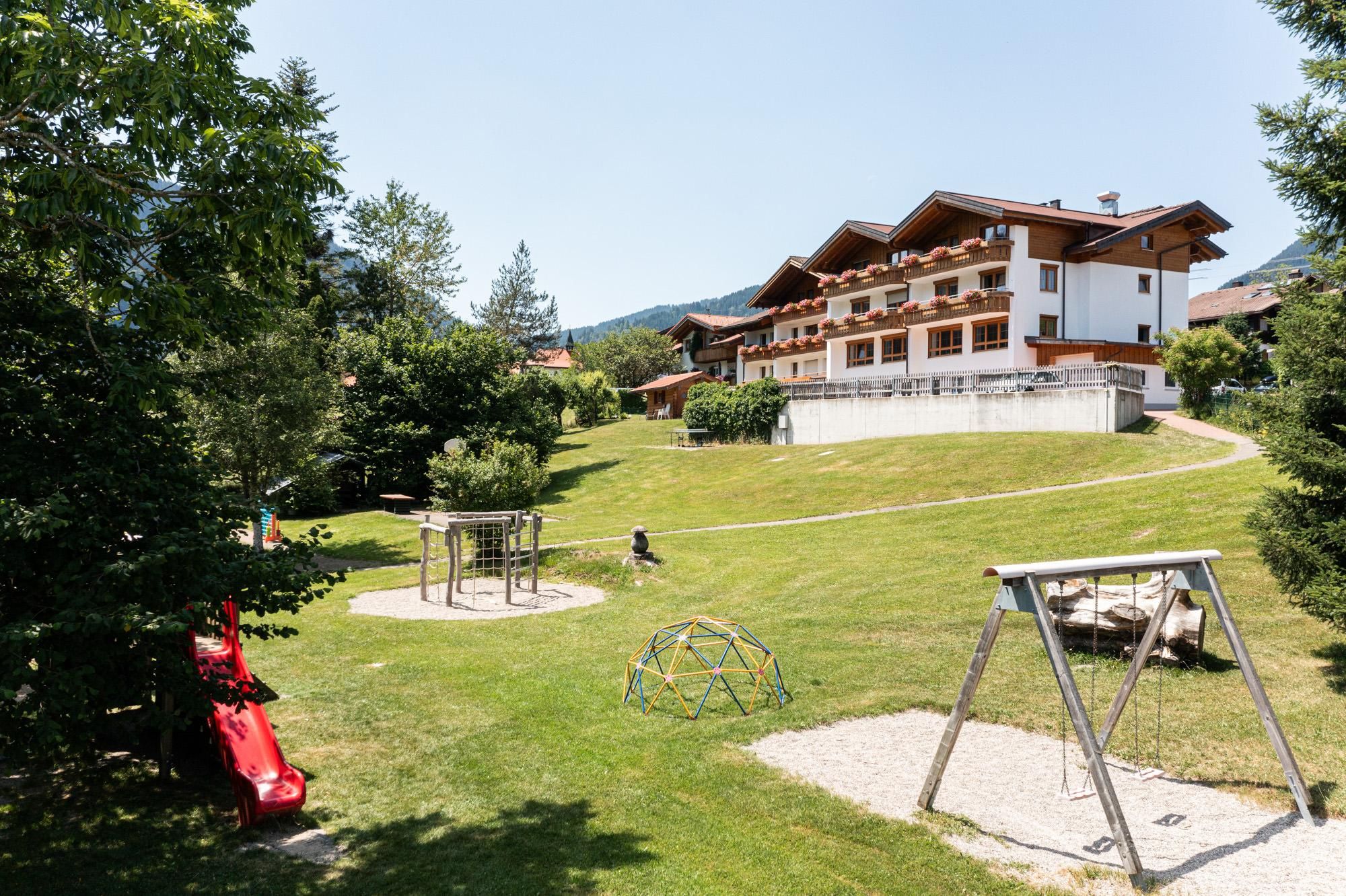 Mutter-Kind-Kurheim: Mehrstöckiges Gebäude mit Balkonen, großer Garten mit Spielplatz, Schaukel, Rutsche, Klettergerüst, Bäume.