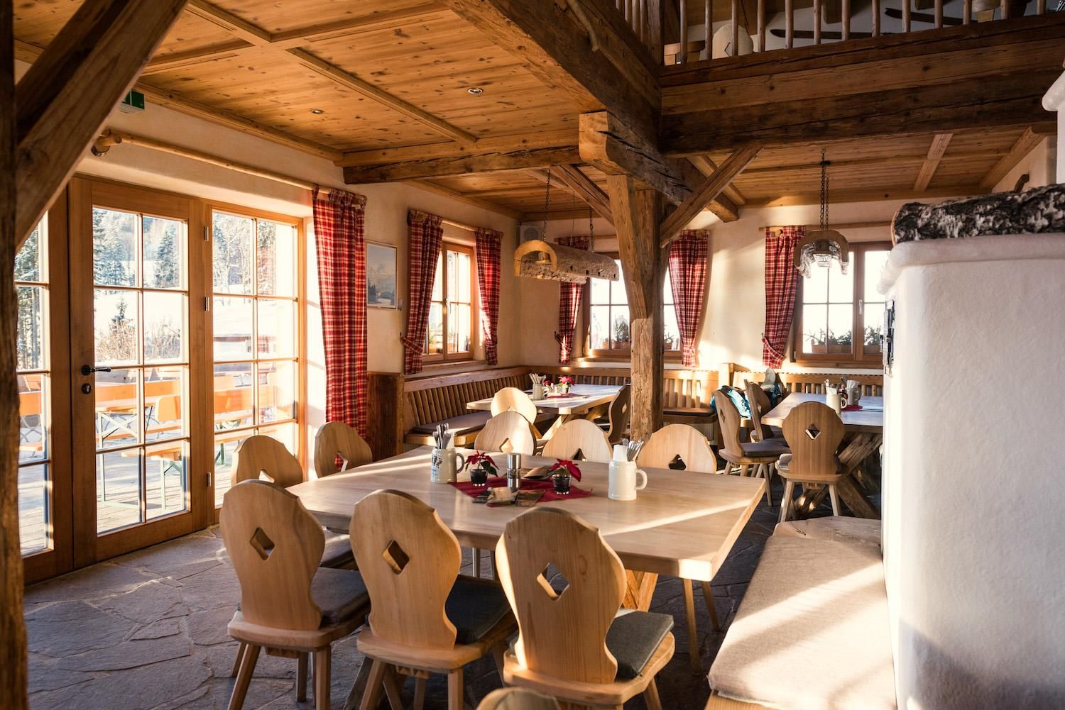 Gemütlicher Gastraum der Schlitte Hitte mit Holzbalkendecke, Holztischen und -stühlen, rot-weiß karierten Vorhängen und einem Kaminofen.