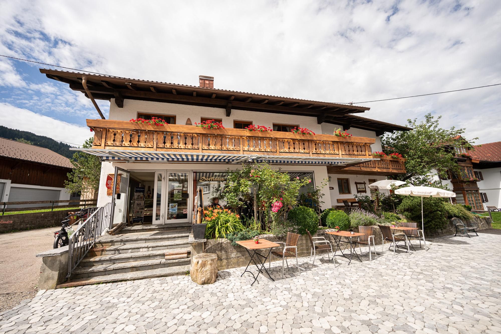 Der charmante Dorfladen in Bolsterlang mit einem blühenden Garten und einer gemütlichen Terrasse. Die Fassade ist traditionell mit Holzverzierungen und blühenden Pflanzen.
