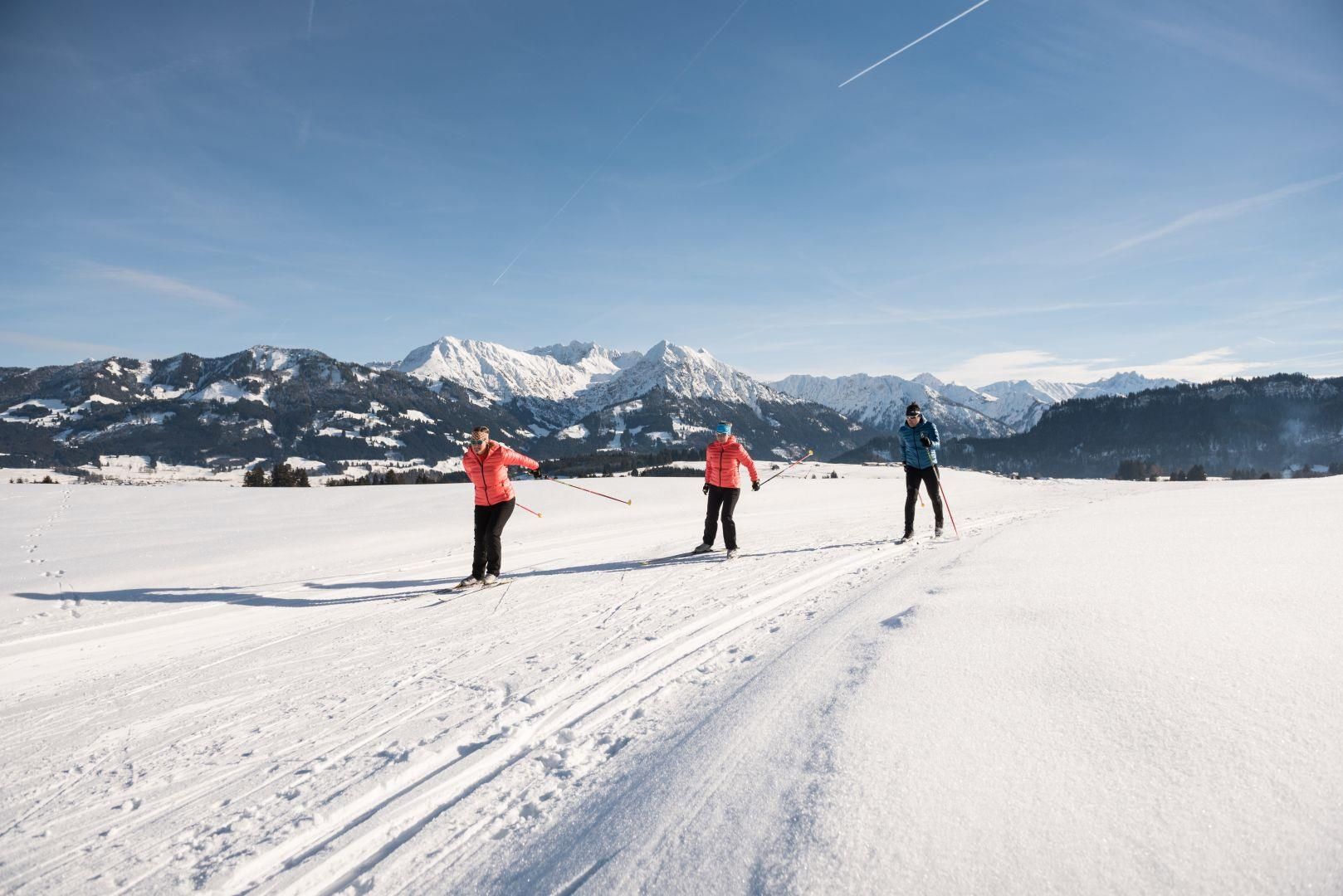 Drei Langläufer auf einer schneebedeckten Loipe mit majestätischen Bergen im Hintergrund.