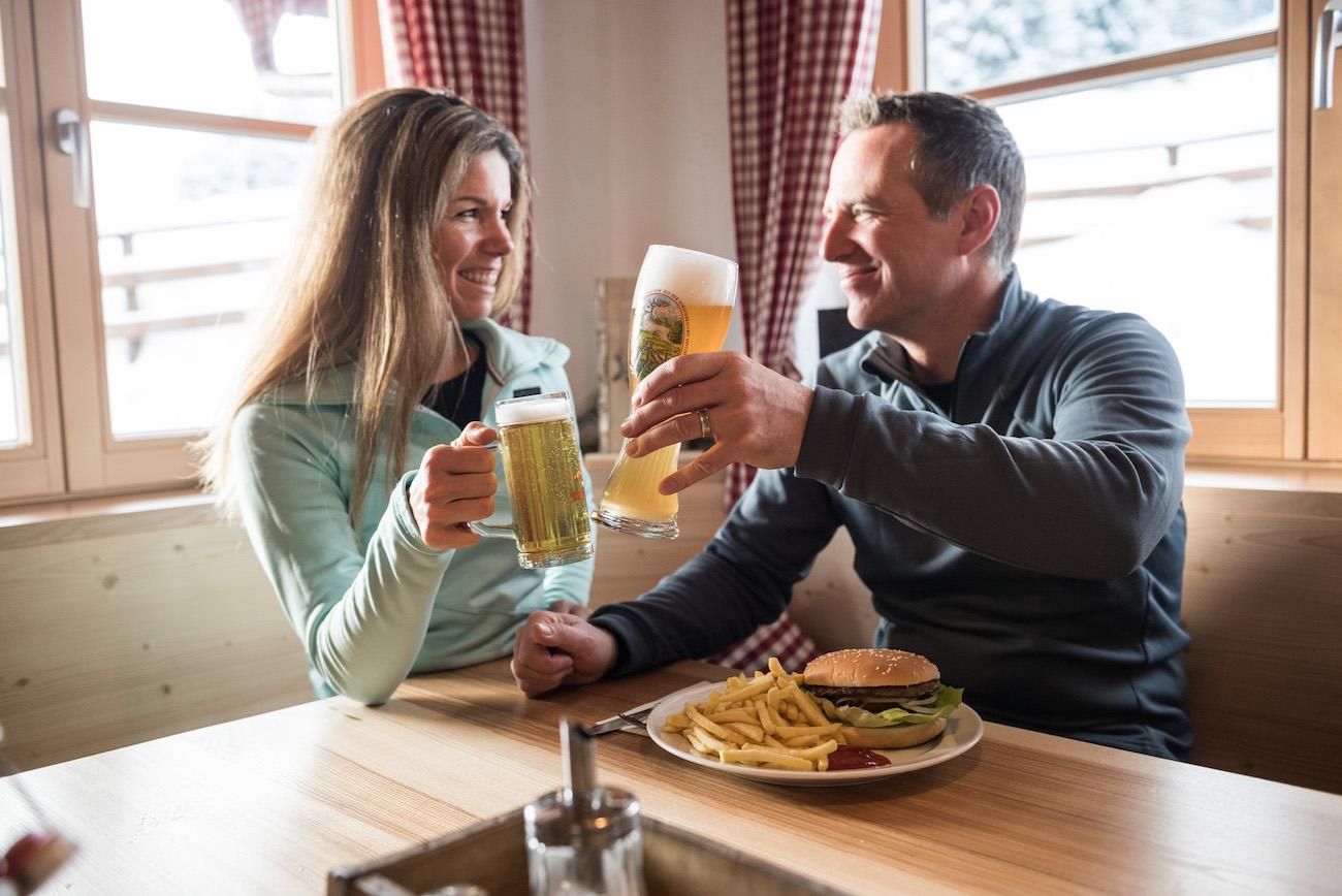 Ein Mann und eine Frau stoßen lächelnd in einem gemütlichen Restaurant auf der Leitenbach Hütte in Ofterschwang miteinander an.