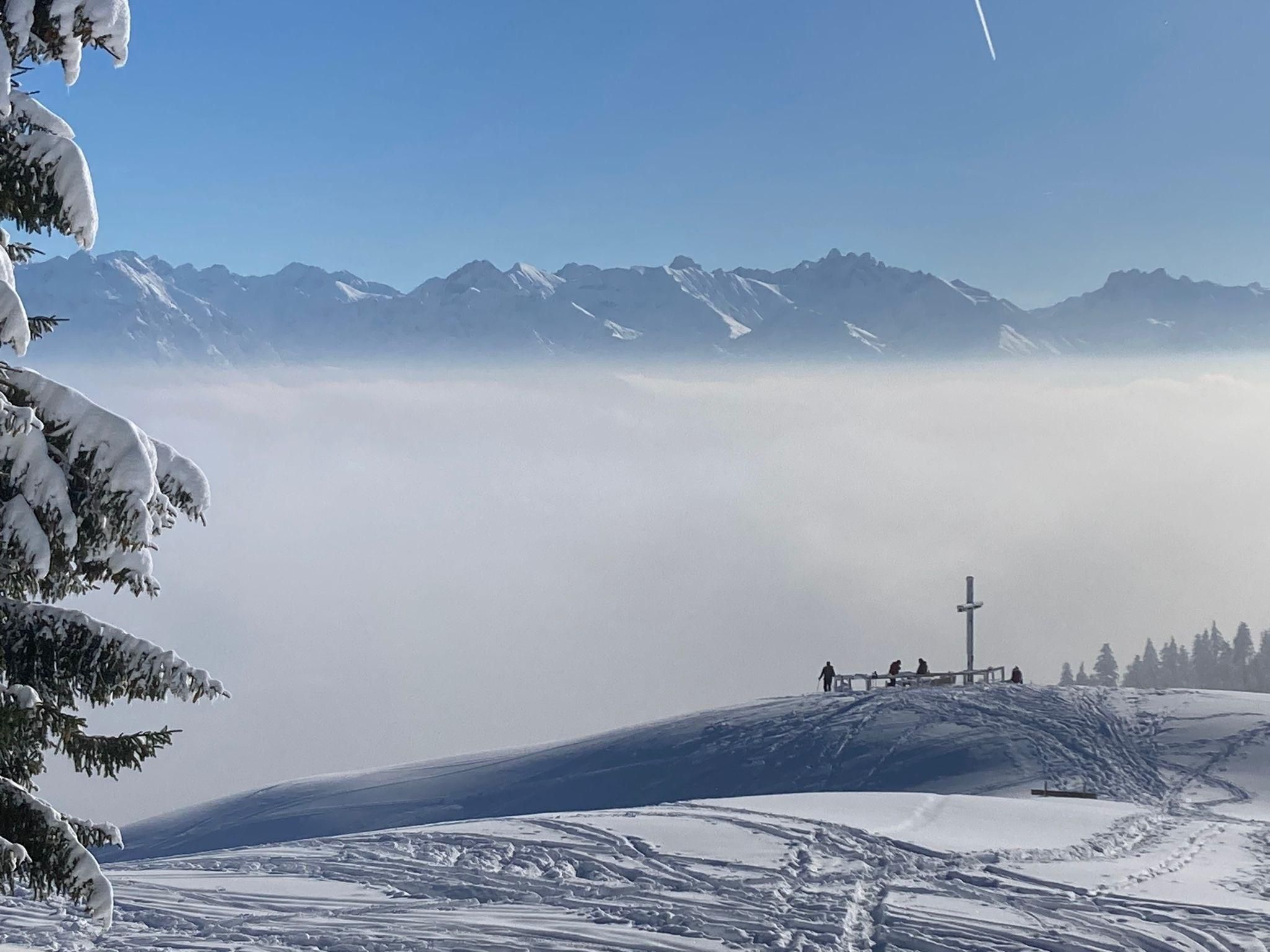 Verschneite Winterlandschaft. Ein Gipfelkreuz auf einem Hügel, umgeben von Skispuren. Eine dichte Nebeldecke liegt im Tal.
