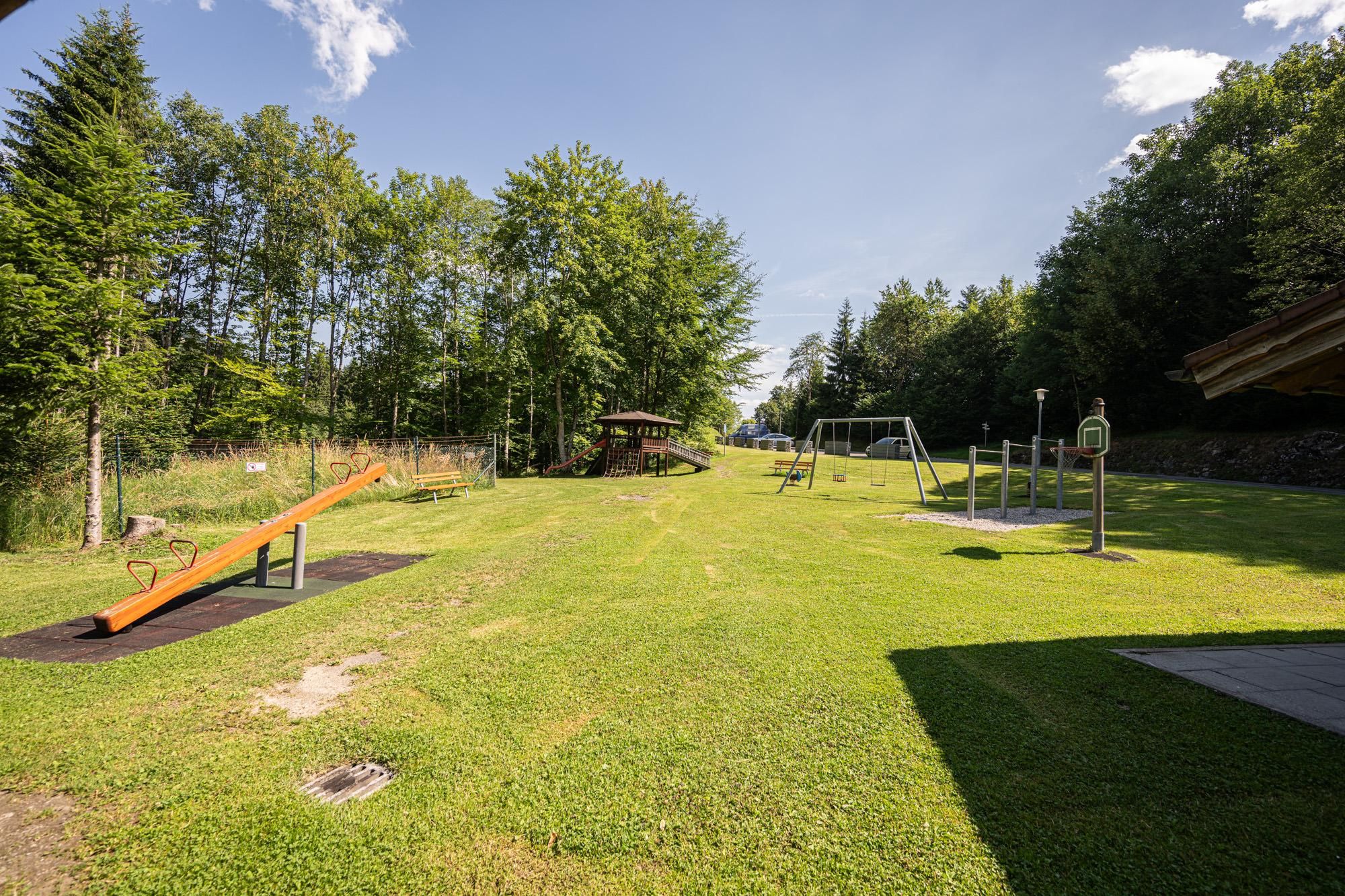 Der Spielplatz am Grillplatz Burgschrofen hat eine Wippe, Schaukeln, ein Klettergerüst mit Rutsche und einen Basketballkorb auf einer grünen Wiese.