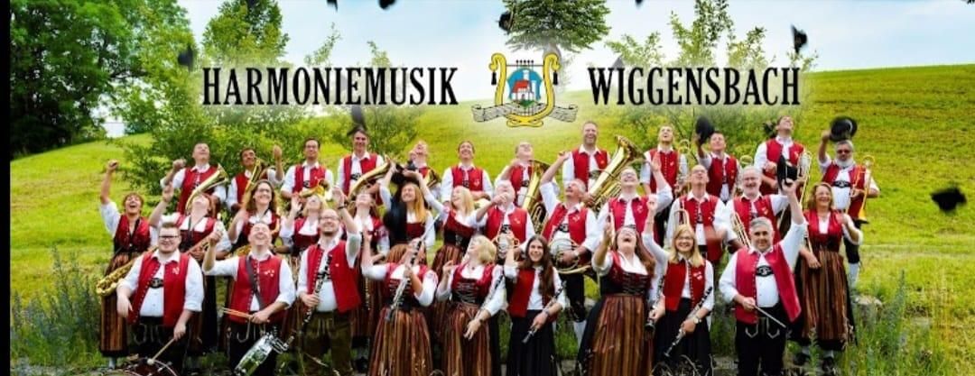 Große Gruppe der Harmoniemusik Wiggensbach in Tracht, jubelnd im Freien. Blasinstrumente und Schlagzeug.