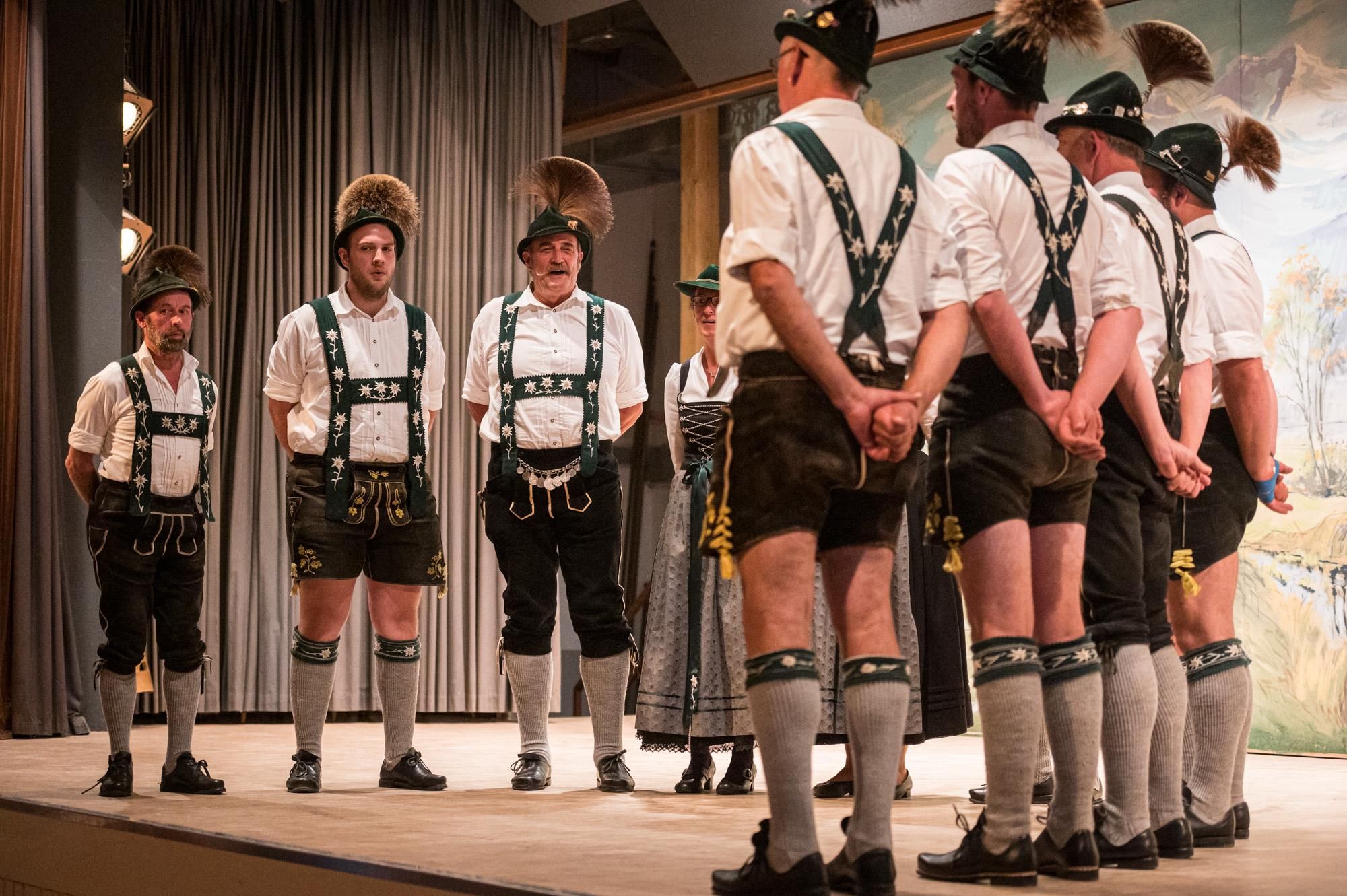Heimatabend: Die Jodlergruppe "Maiselstuinar" in Tracht auf der Bühne. Hinten eine Frau im Dirndl und ein Mann, der sich die Infotafel des Themenwegs anschaut.